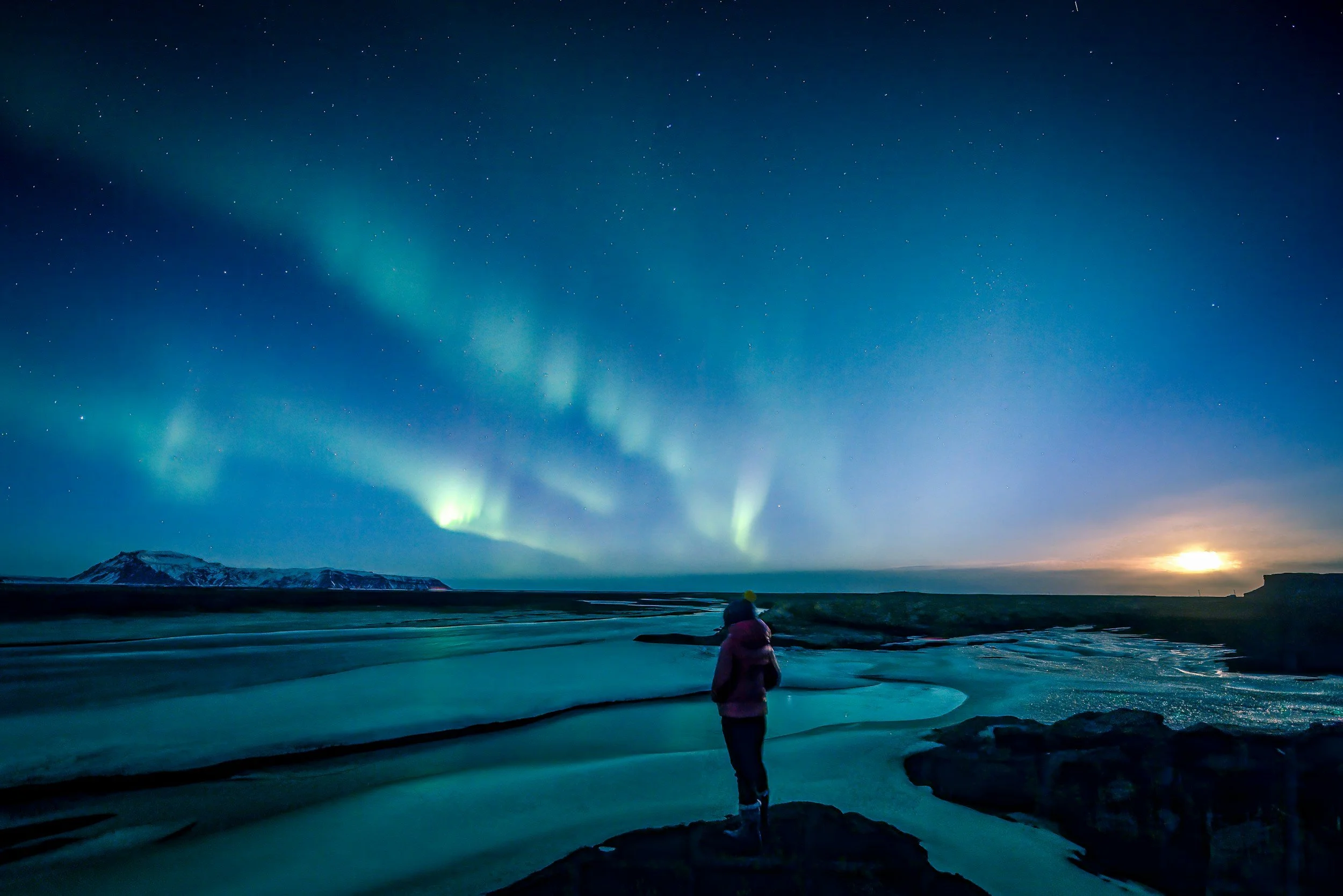 A person standing on a rocky outcrop watching the Northern Lights illuminate the night sky over a frozen landscape.