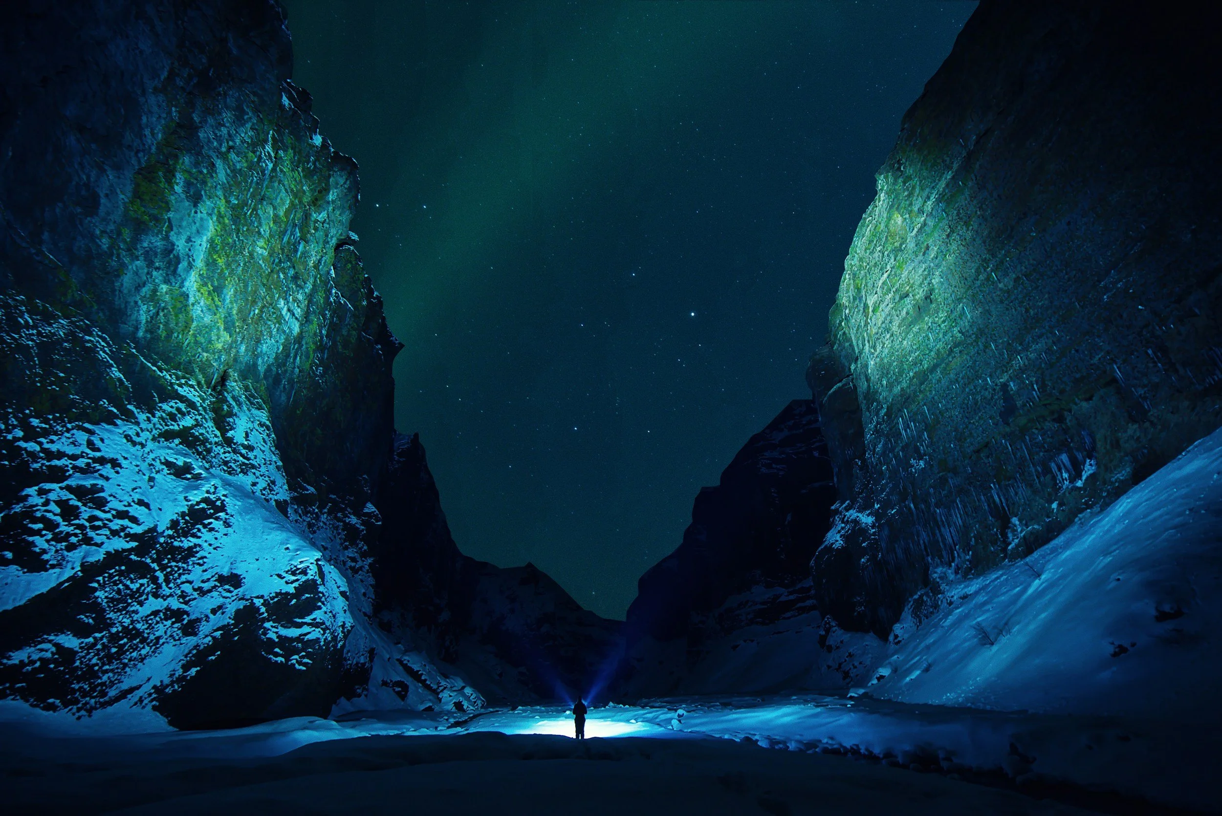 A person standing in a snow-covered canyon at night, illuminated by a flashlight, with the northern lights and stars in the sky.