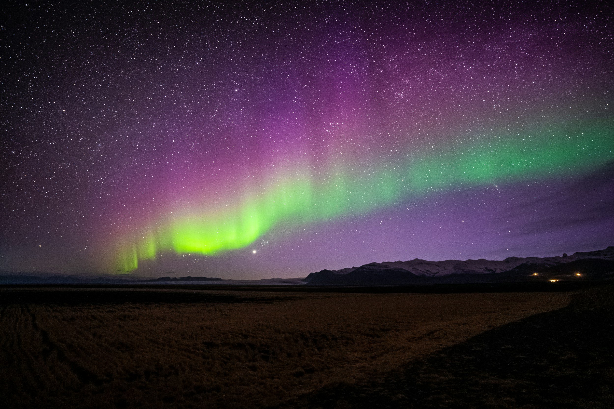 Northern lights in the night sky with stars over a mountainous landscape and sandy foreground.