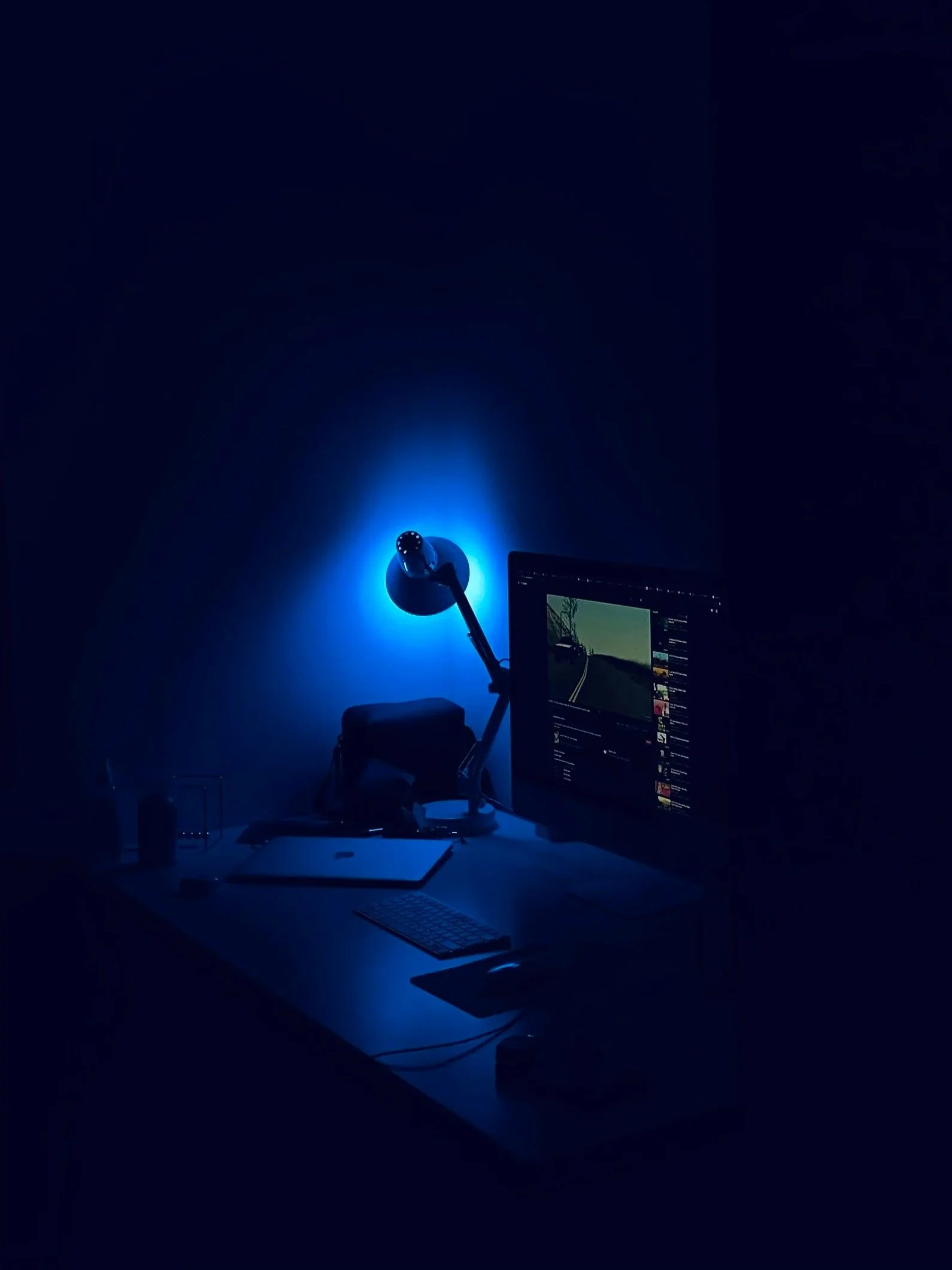 A dark room with a desk illuminated by a blue light. The desk has a computer monitor displaying a video, a closed laptop, a wireless keyboard, a mouse, and a desk lamp.