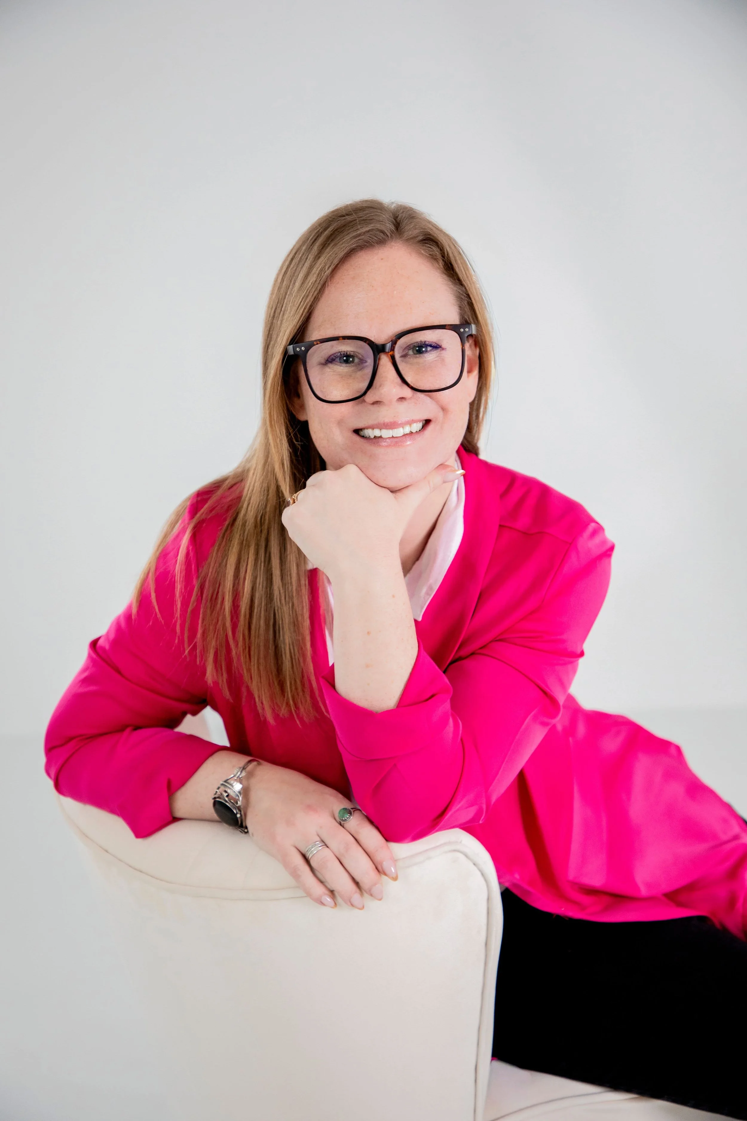 Woman with red hair and glasses smiling, wearing a pink jacket, sitting on a white chair against a white background.