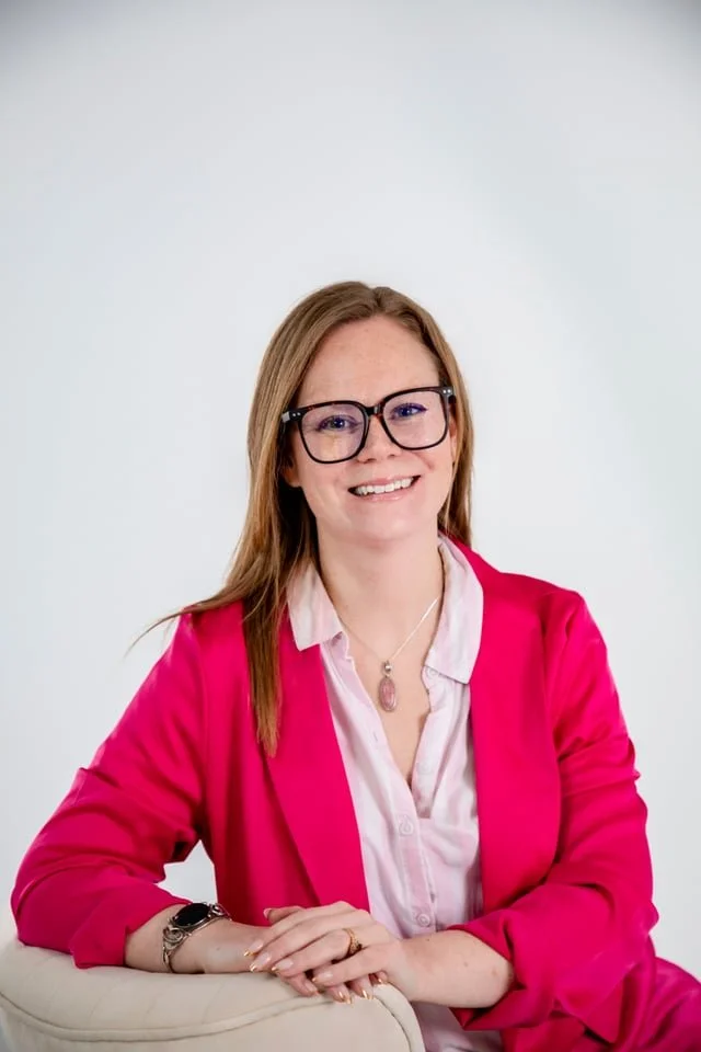 A woman with long brown hair, wearing glasses, a pink blazer, and a white shirt, sitting with her hands folded on a white chair, smiling at the camera against a plain white background.