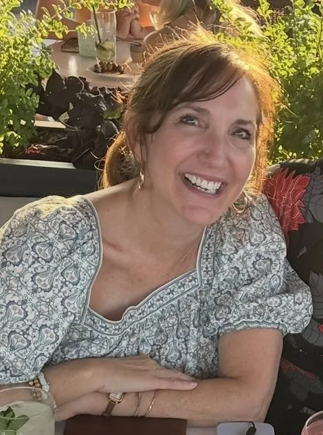 A smiling woman with brown hair, wearing a patterned top, sitting outdoors at a table during sunset with greenery around.