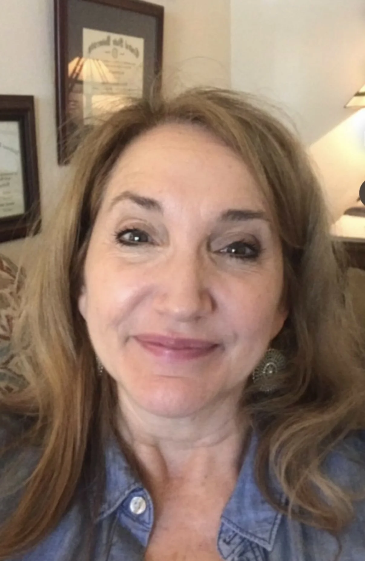 A woman with shoulder-length curly hair smiling at the camera. She is wearing earrings and a denim shirt. In the background, there are framed certificates or diplomas on the wall and a lamp.