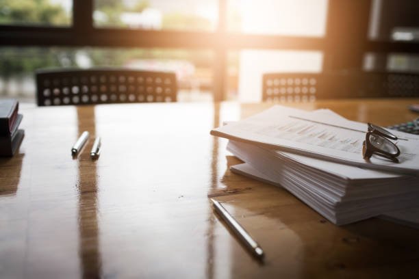 A wooden conference table with stacked papers, eyeglasses, and pens in front of large windows with sunlight.
