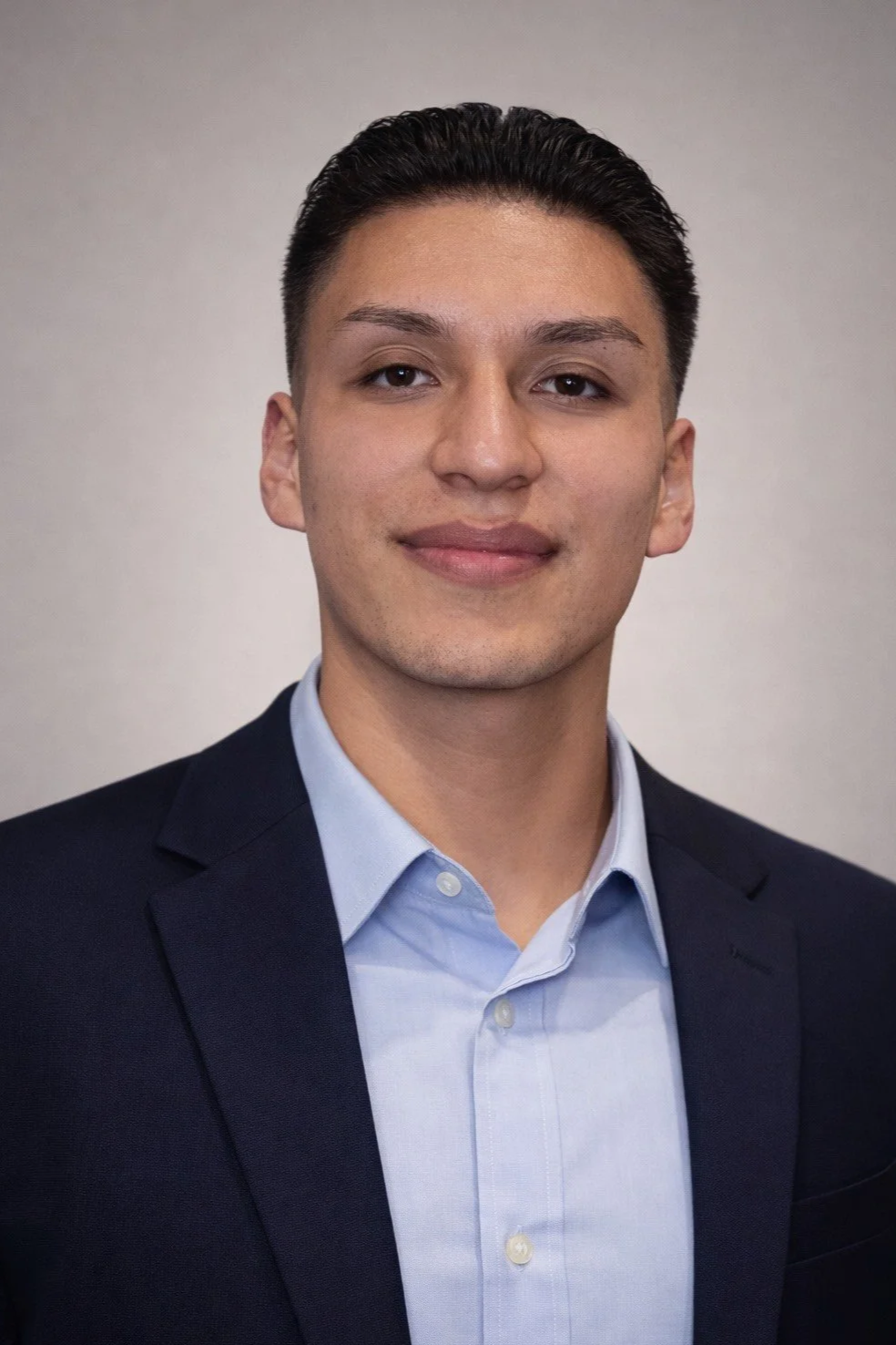 Headshot of a young man with dark hair, wearing a blue dress shirt and a dark blazer, against a neutral background.