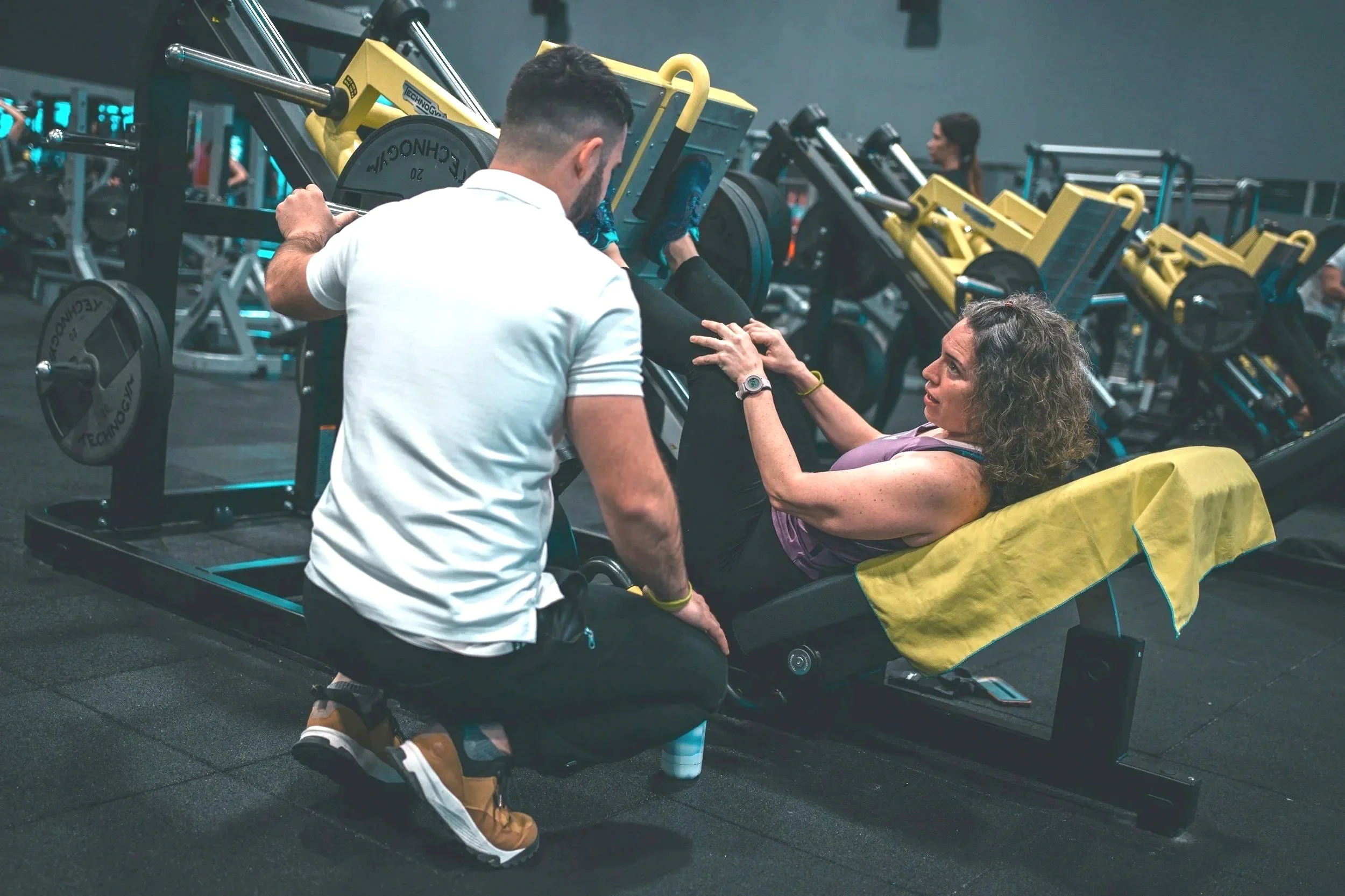 A woman is lying on a leg press machine in a gym while a trainer assists her with her workout, with gym equipment and other people in the background.