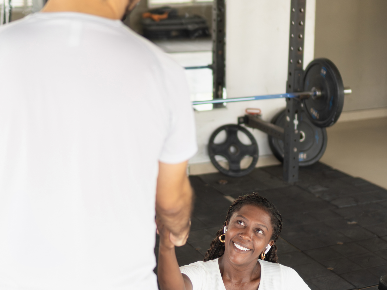 A woman with braids and earrings smiling while working out in a gym, shaking hands with a man in a white shirt.