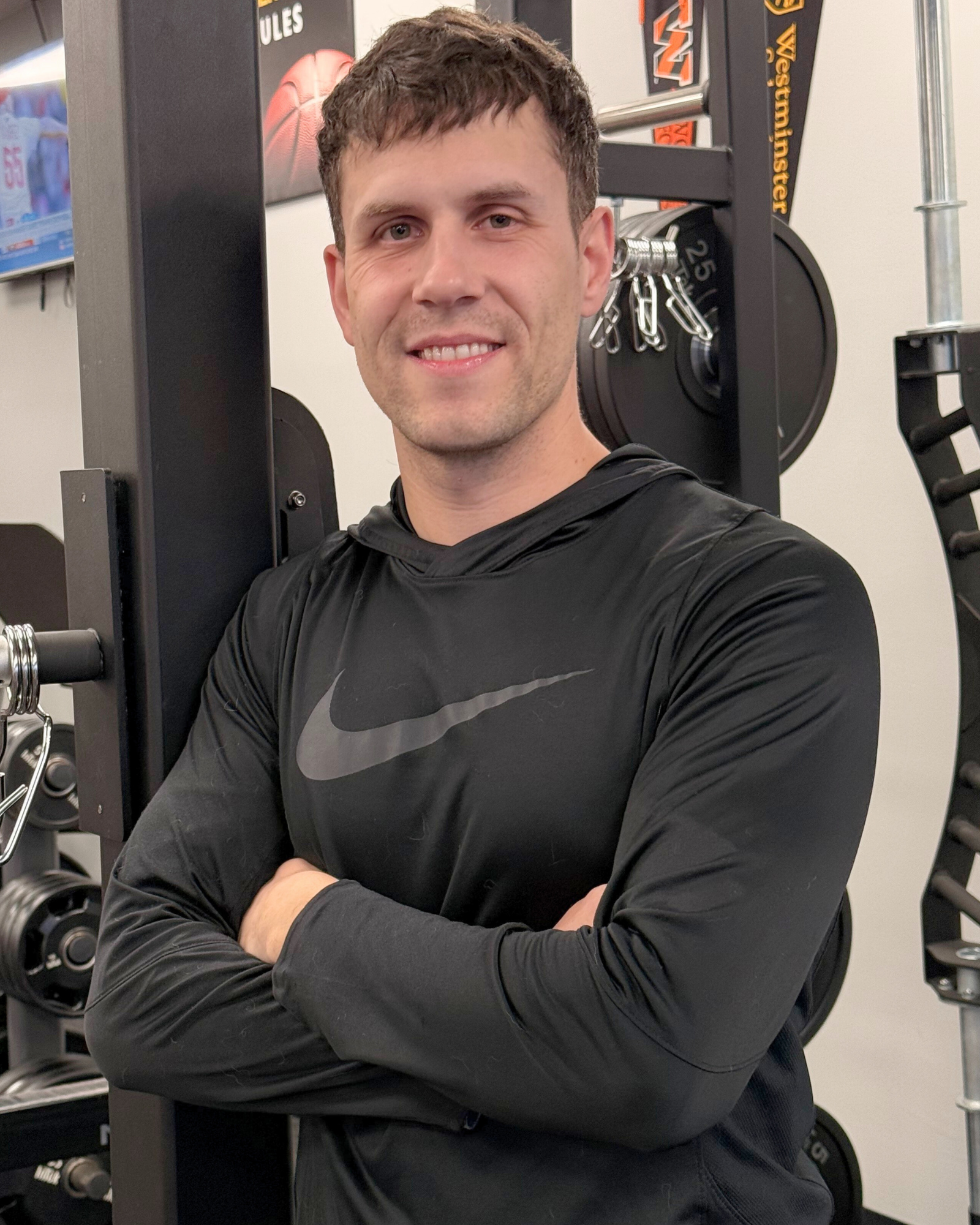 A young man in a black Nike hoodie standing with arms crossed, smiling at the camera in a gym with weightlifting equipment and motivational posters on the wall.