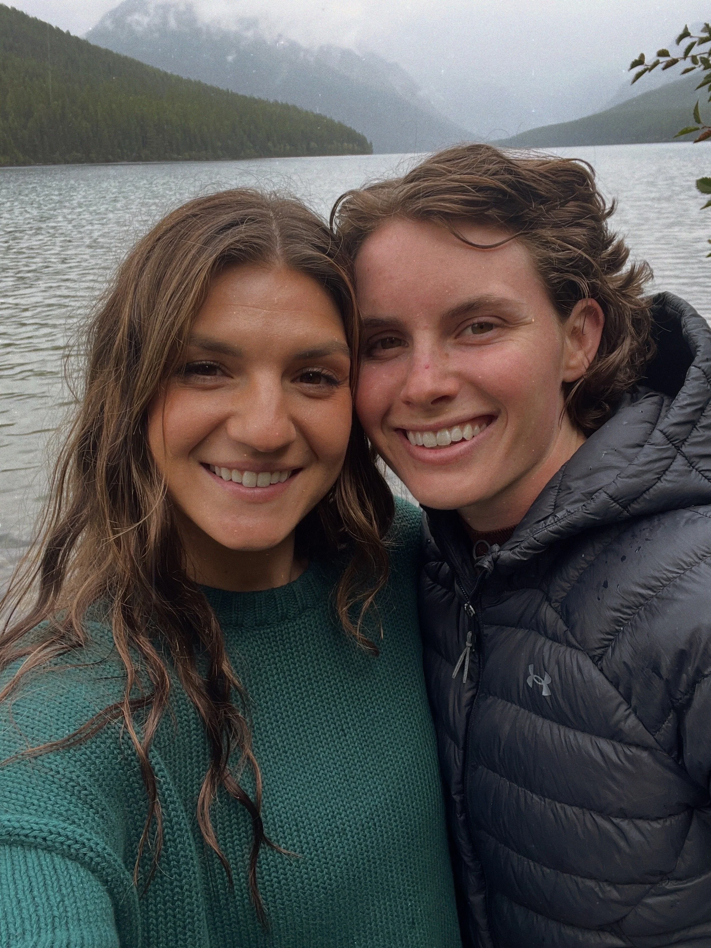 Two smiling young adults taking a selfie outdoors near a lake with mountains in the background.