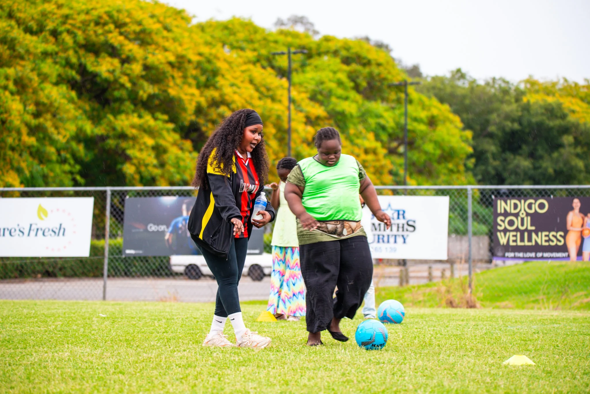 Two women, one wearing a black and red sports jersey and the other in a green and brown shirt, are engaging in a soccer activity on a grassy field with two soccer balls. There are trees and advertising banners in the background.