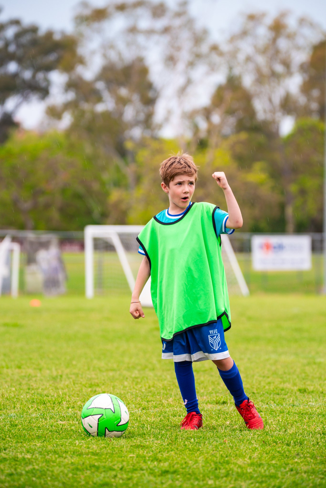 A young boy on a soccer field wearing a blue uniform and a green training bib, standing next to a soccer ball with a colorful pattern, with a soccer goal and trees in the background.