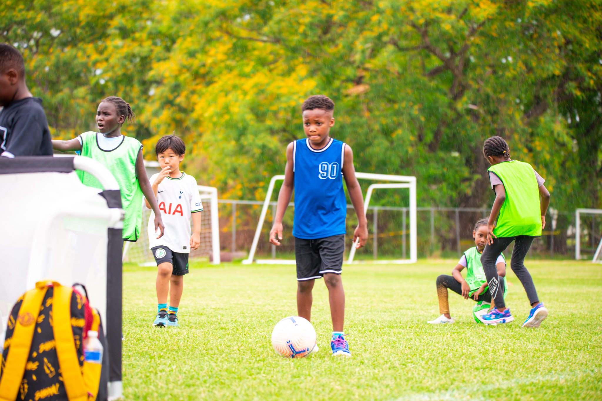 Children playing soccer on a field during daytime, with trees in the background.