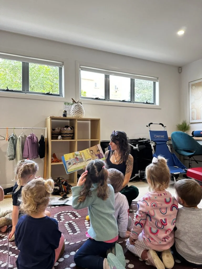 A woman sitting on the floor reading a children's book to six young kids in a classroom or daycare setting. The kids are sitting on a patterned rug, paying attention. The room has large windows, a bookshelf, and some furniture in the background.