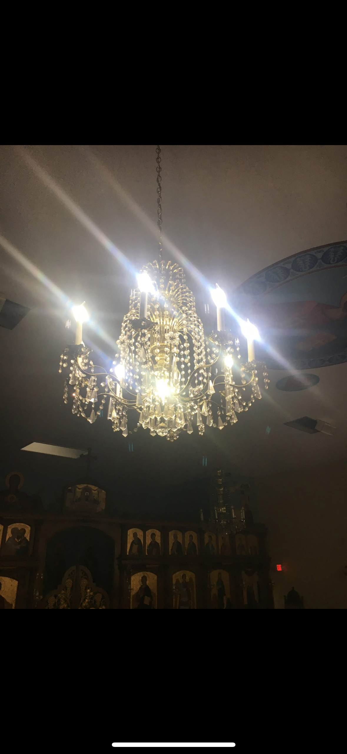 A chandelier with multiple lights and hanging crystals illuminated against a ceiling in a dimly lit room, with religious icons and artwork in the background.