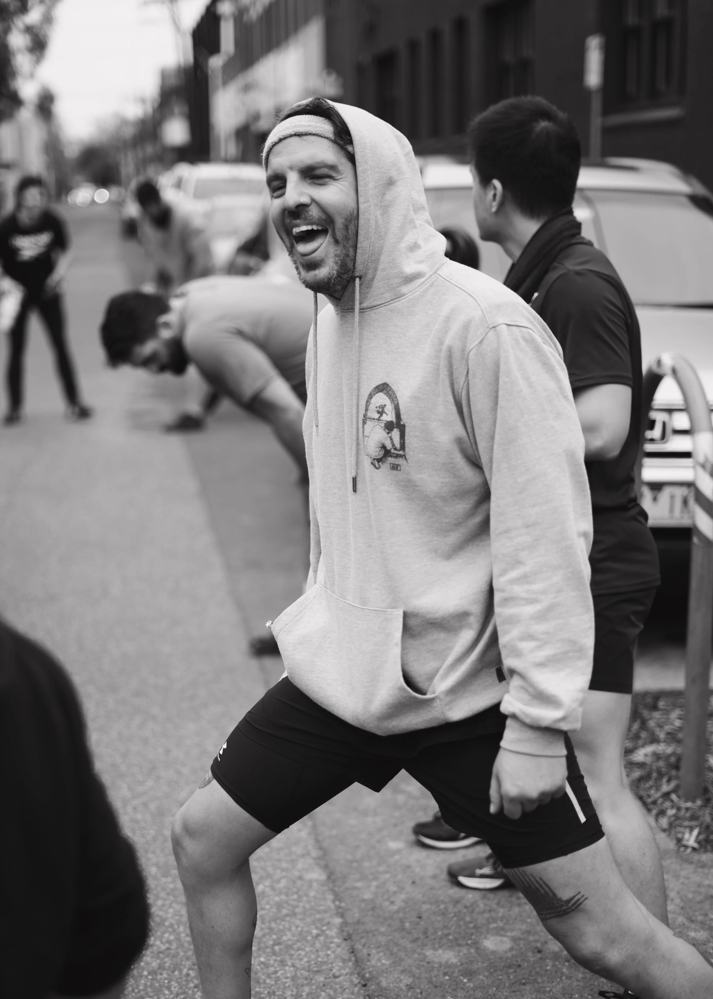 A man in a hoodie and shorts smiling outside with other people stretching or warming up in the background.