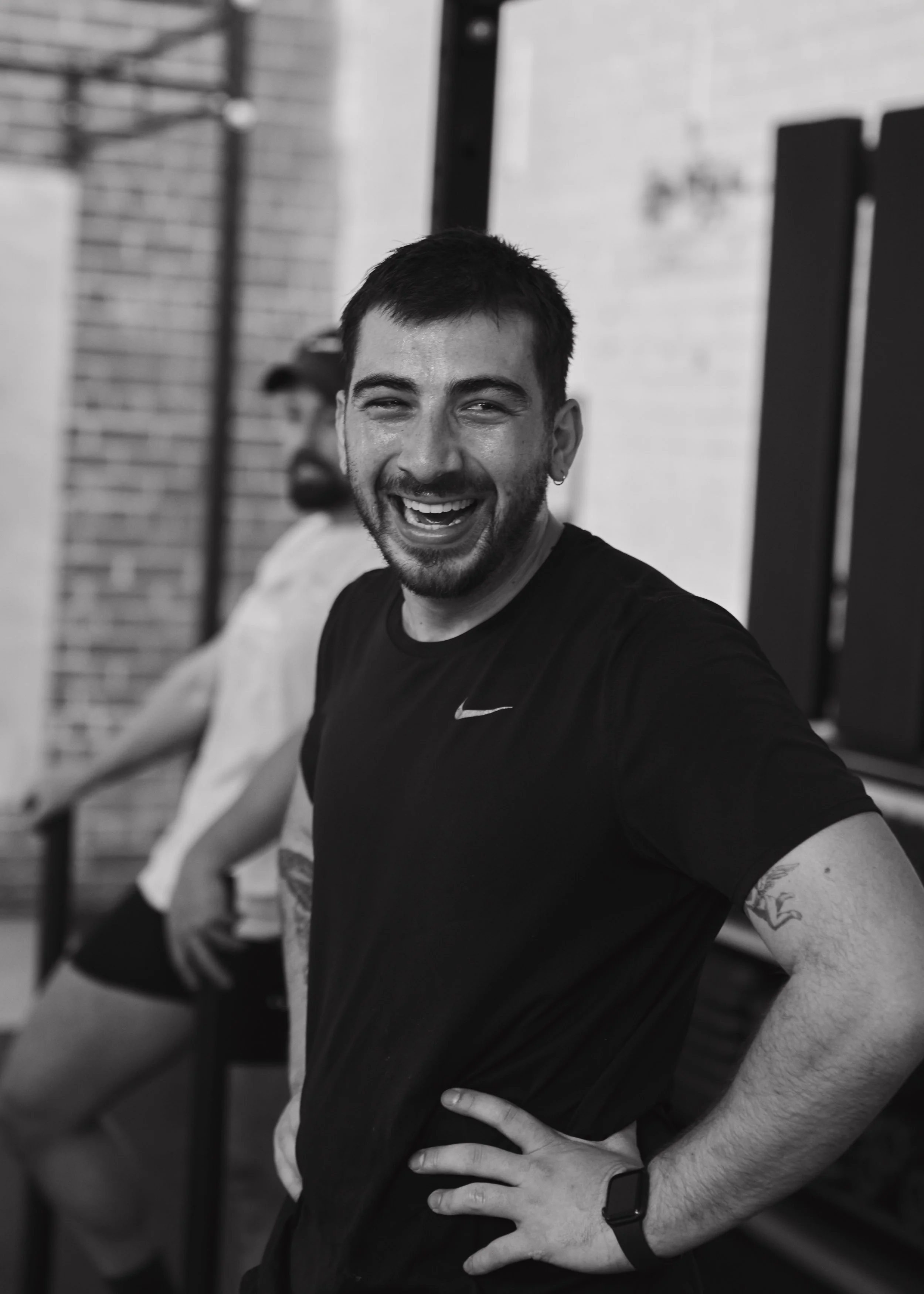 A smiling man with short dark hair and a beard, wearing a black Nike t-shirt and a smartwatch, standing with his hand on his hip in a gym setting.