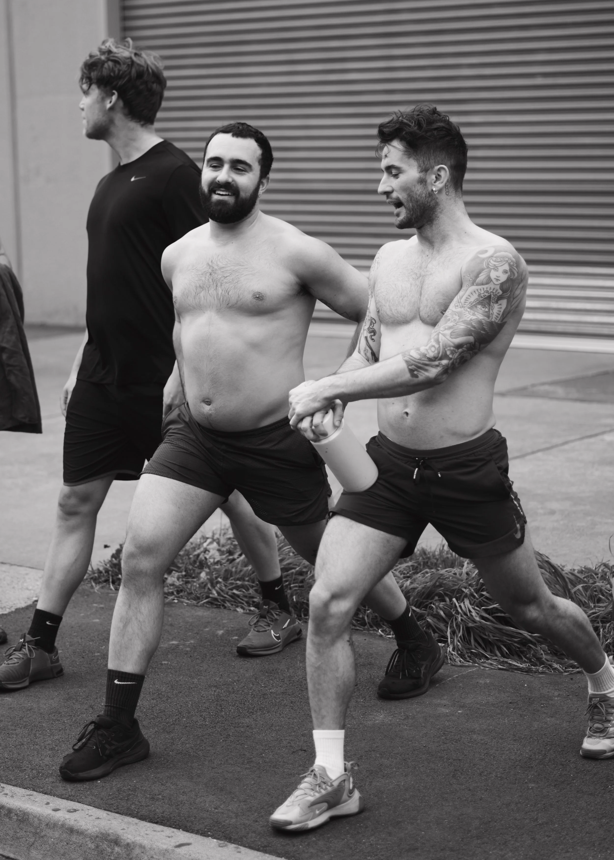 Three shirtless men in shorts outdoors, one holding a water bottle, walking on a sidewalk near a building with a metal shutter. Two of the men are smiling, while the third man in the background is turned away.