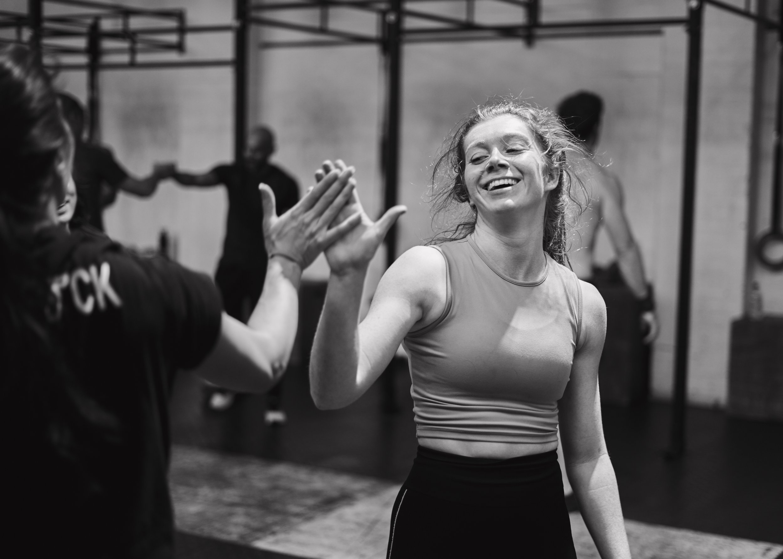 A woman smiling and high-fiving a trainer in a gym, with other people working out in the background.