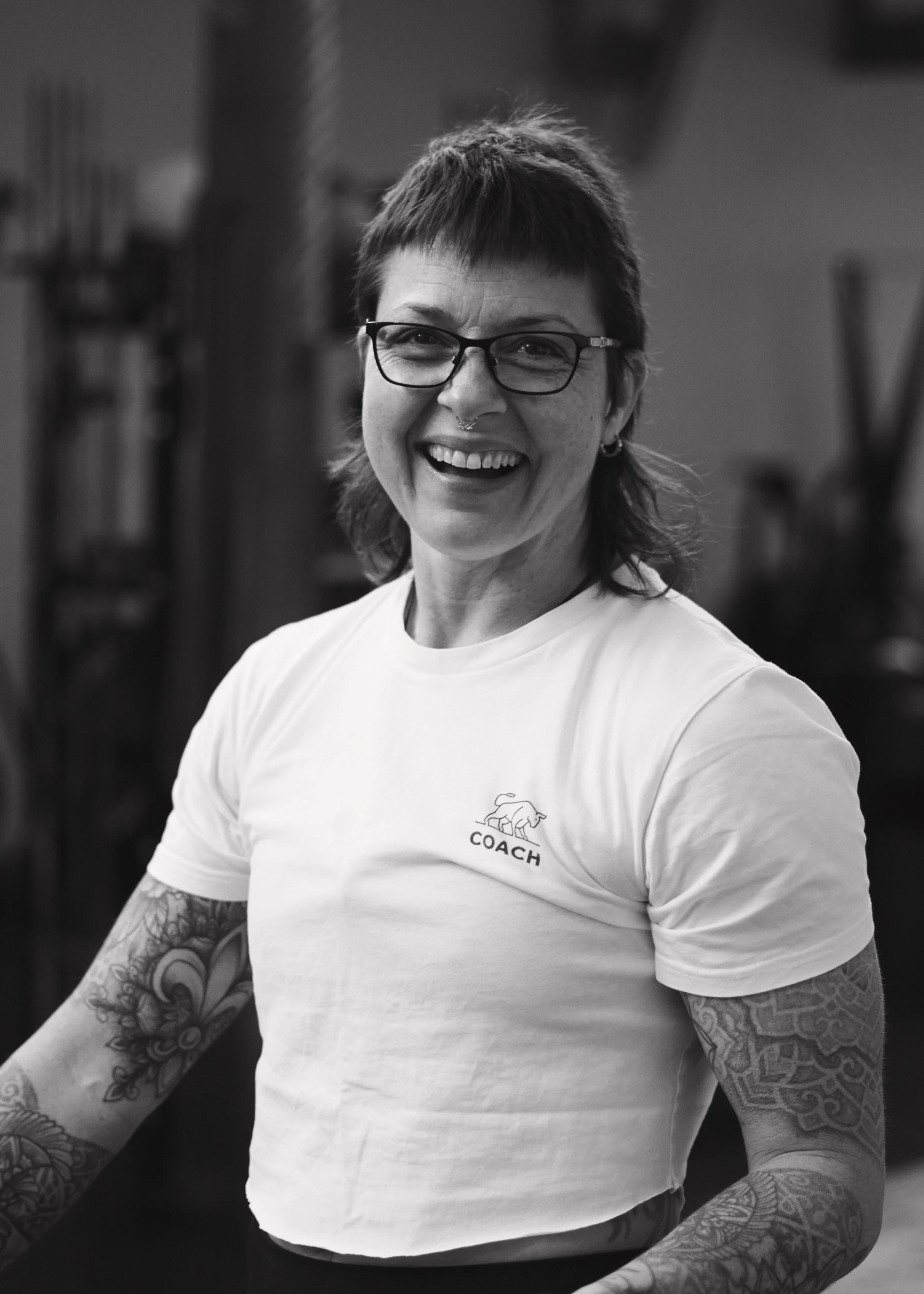 A smiling woman with short hair, glasses, and tattoos on her arms, wearing a white T-shirt with a logo and the word 'COACH', in a gym environment.