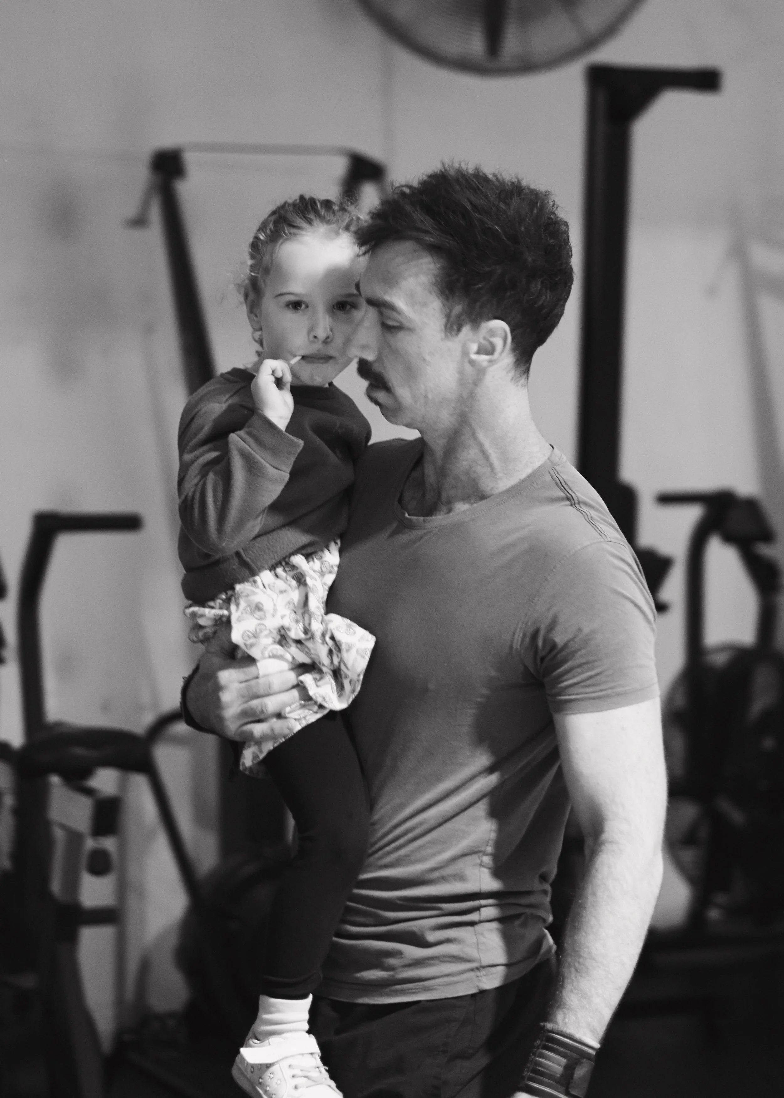 A man holding a young girl in his arms in what appears to be a gym. The man is wearing a T-shirt and the girl is in a sweater and leggings. They are close together, and the girl is touching her face with her finger. The background has gym equipment a
