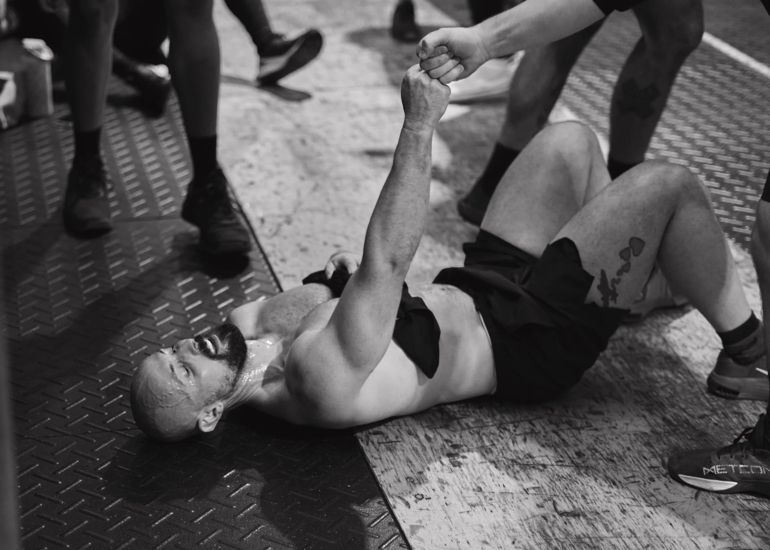 A man with a beard lying on the ground, shirtless, doing a one-arm push-up while holding hands with another person standing above him. The scene appears to be at a gym or fitness event, with other people and gym equipment visible around them.