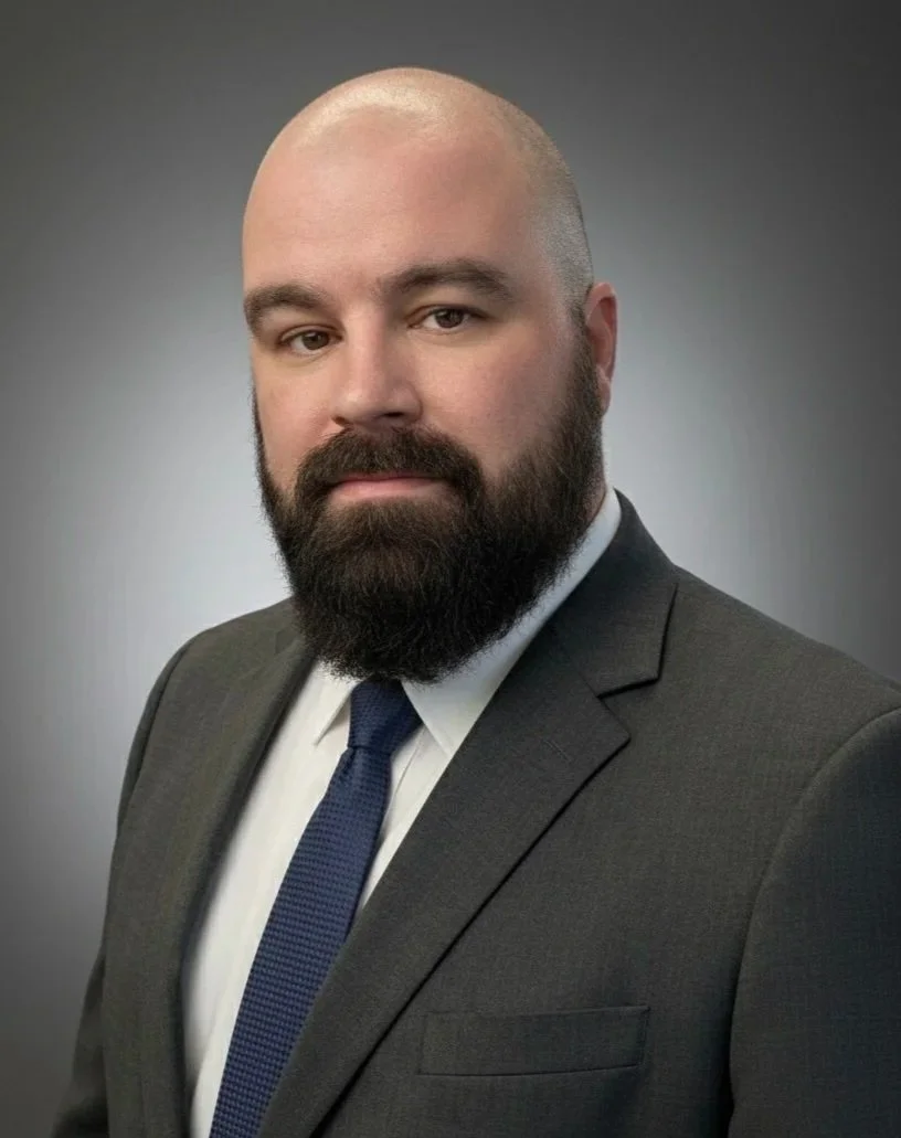 Portrait of Michael Clarkson in a charcoal suit, blue tie, and white shirt. Michael has a shaved head and a neatly trimmed beard.