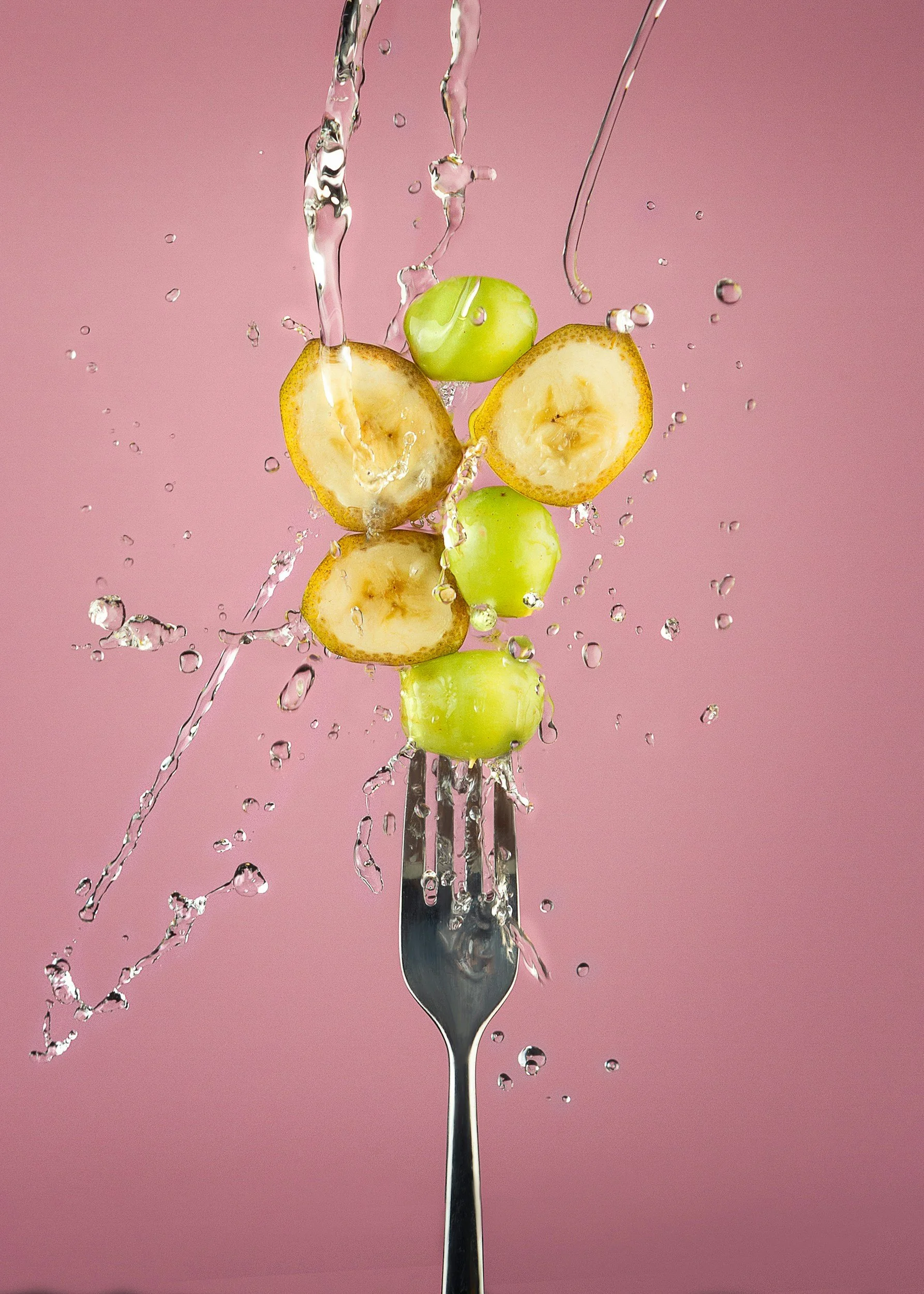 Green grapes and banana slices on a fork with water splashing against a pink background.