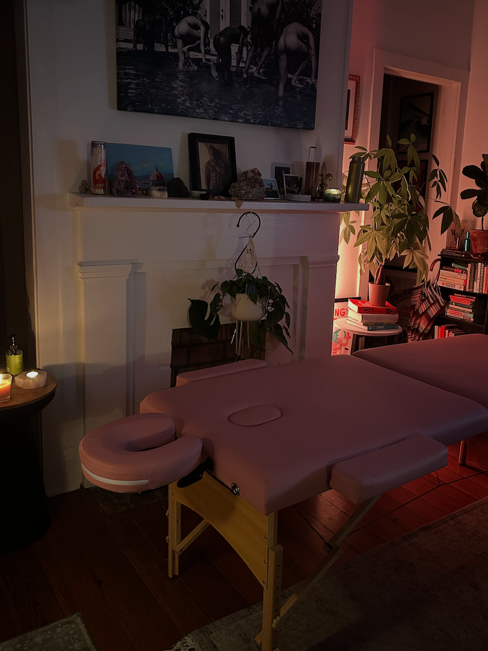 A massage table in a cozy, dimly lit room decorated with plants, framed pictures, and a large black-and-white photograph of people swimming hung above a white fireplace mantel with various decorative items.