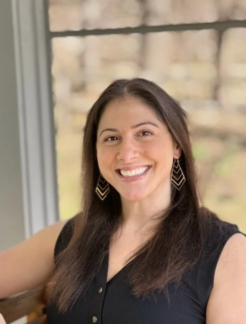 A woman with long dark hair smiling, wearing a black sleeveless top and geometric earrings, seated near a window with a blurred outdoor background.