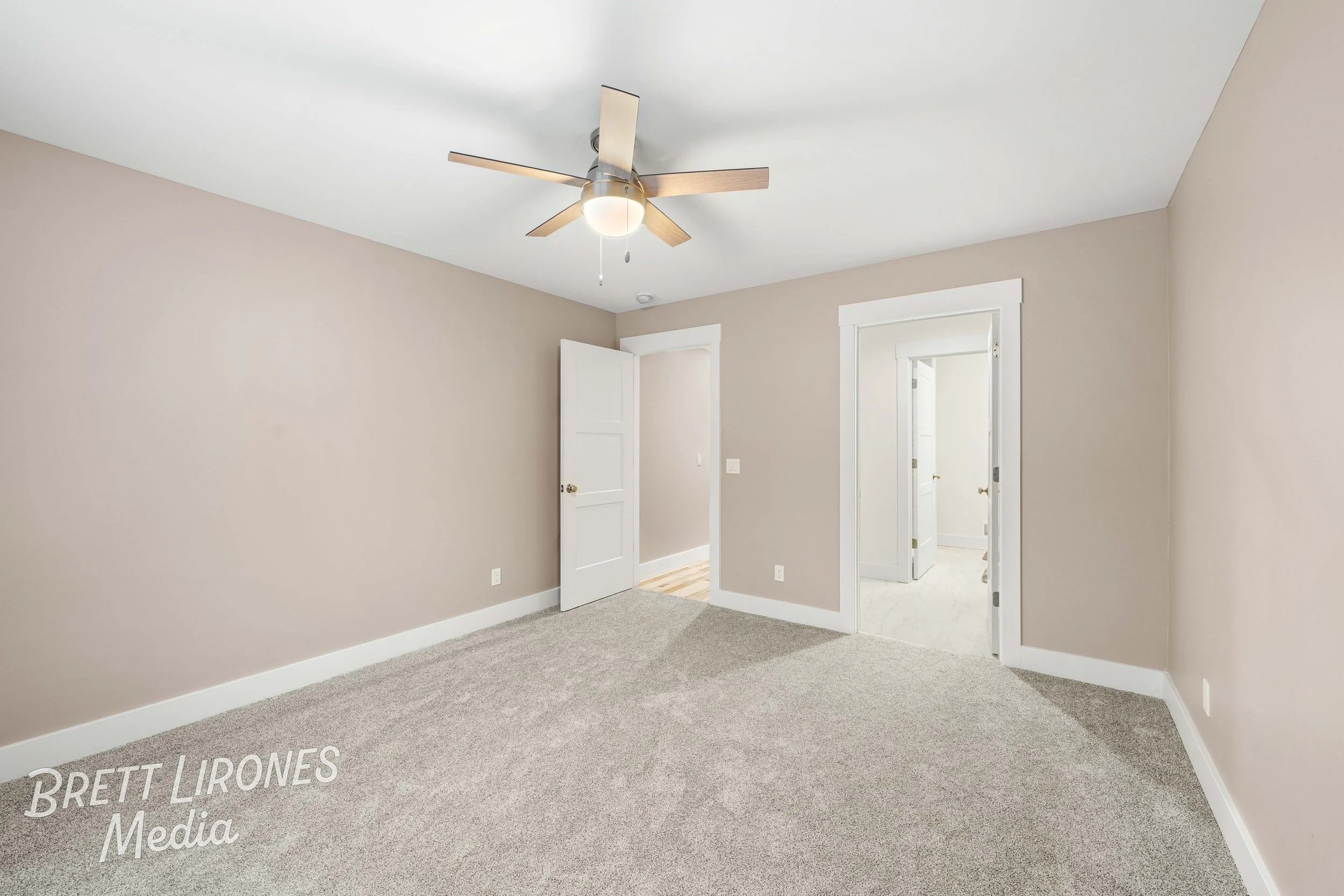Empty beige-colored bedroom with white trim, carpeted floor, ceiling fan, and two open doors leading to adjacent rooms.
