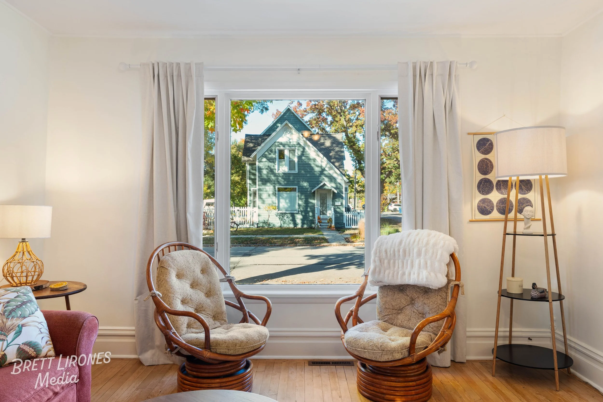 Living room with beige chairs, a side table with a lamp, a large window showing a green house outside, and curtains.