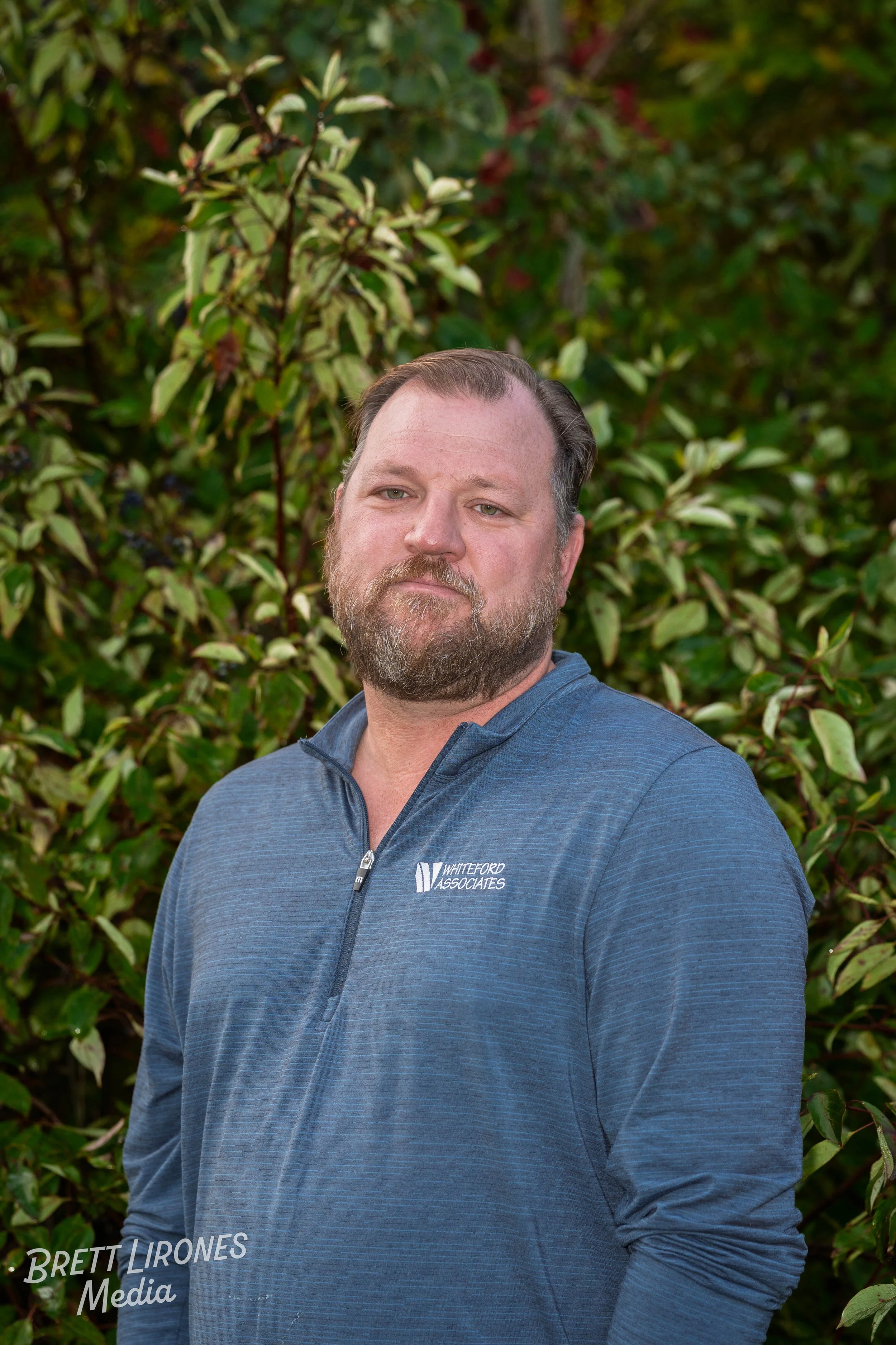 A man with a beard and mustache wearing a blue quarter-zip jacket standing outdoors in front of green foliage.