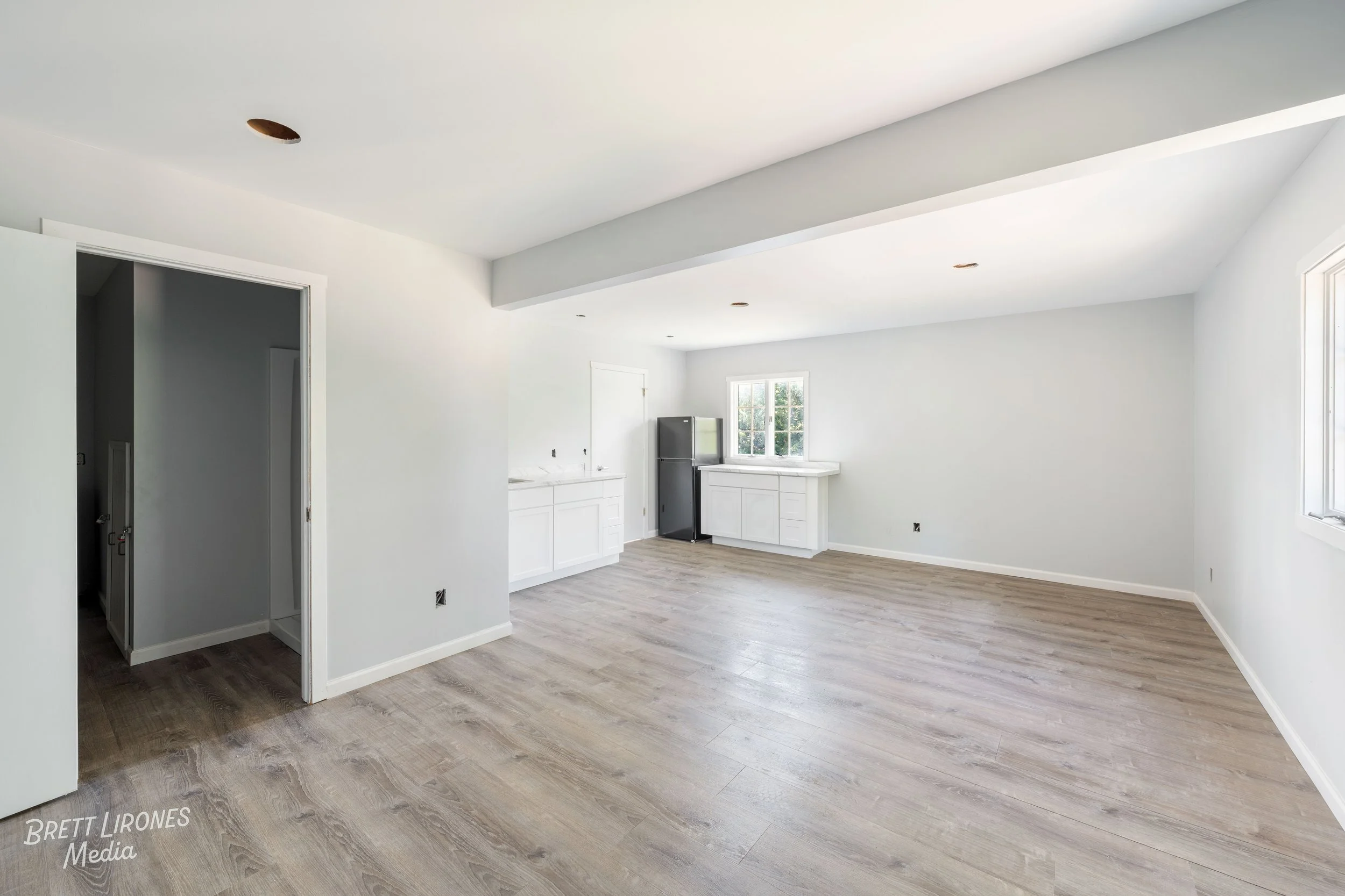 Empty living room and kitchen area with white walls, light wood flooring, and a window letting in natural light, new construction or renovation