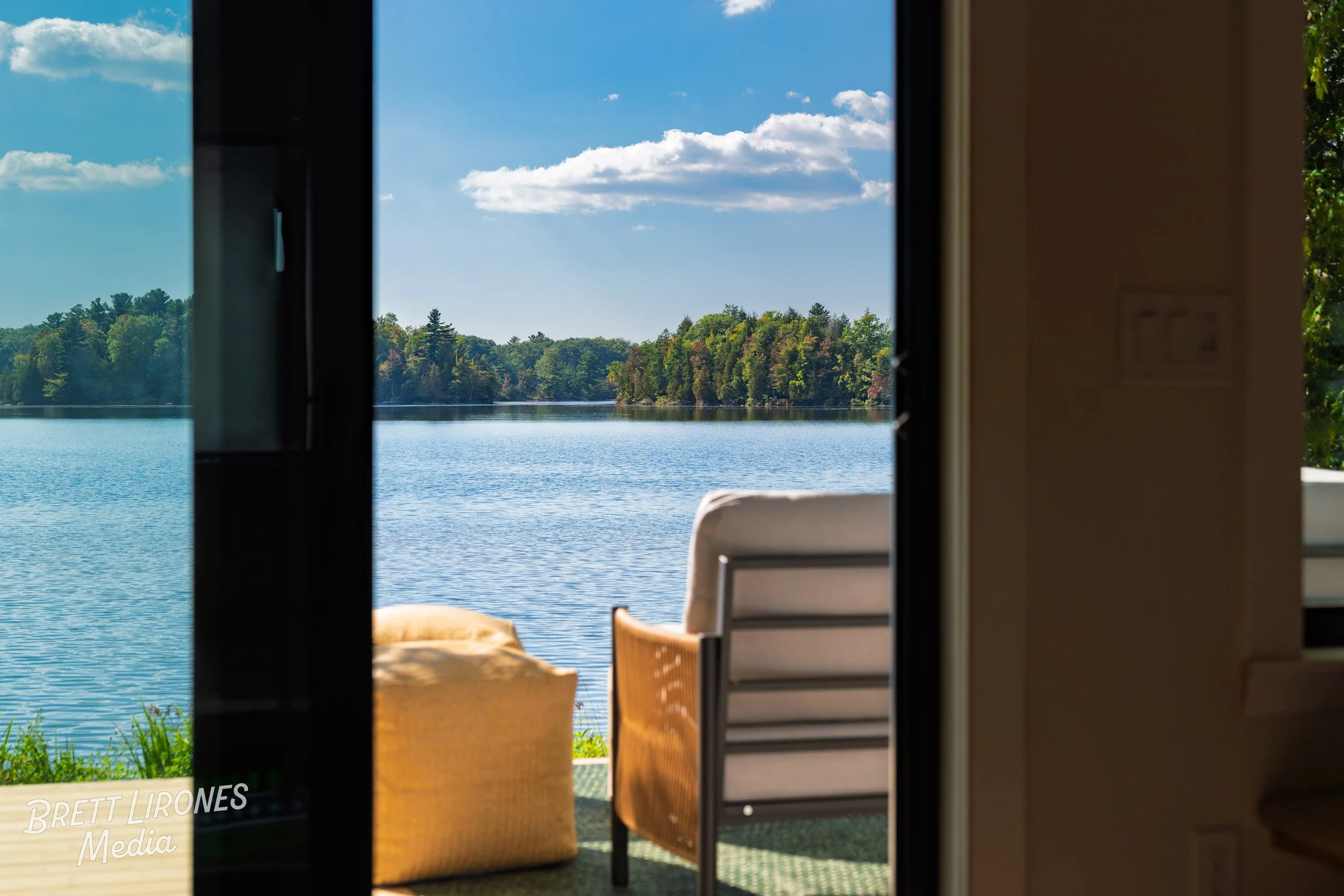 View of a peaceful lake with a tree-lined shore under a blue sky with white clouds, seen through an open door from inside a room with a patio chair and a beige armchair.