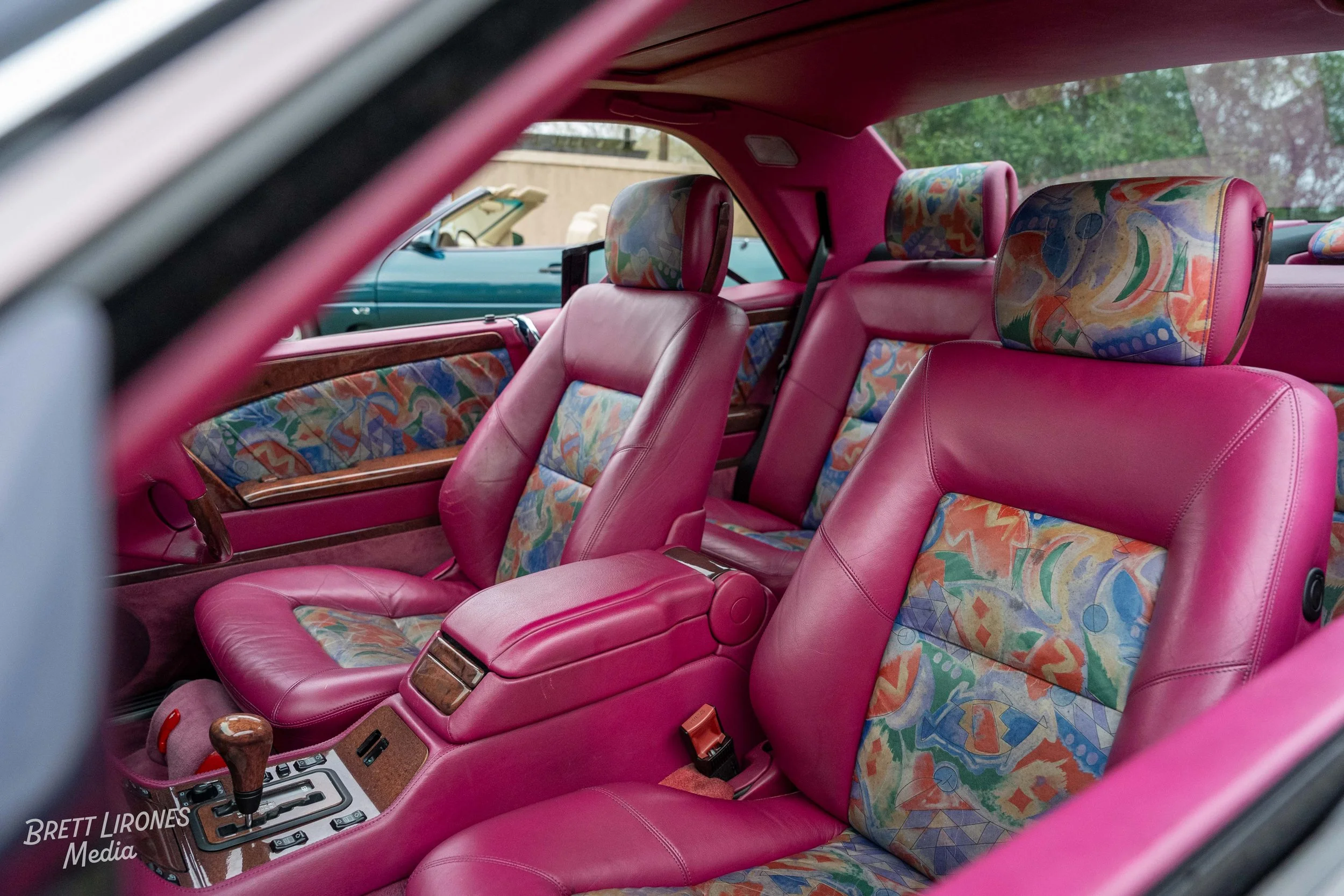 Interior of a vintage car with pink leather seats, colorful patterned fabric on the headrests and seat backs, and a wood-trimmed center console.