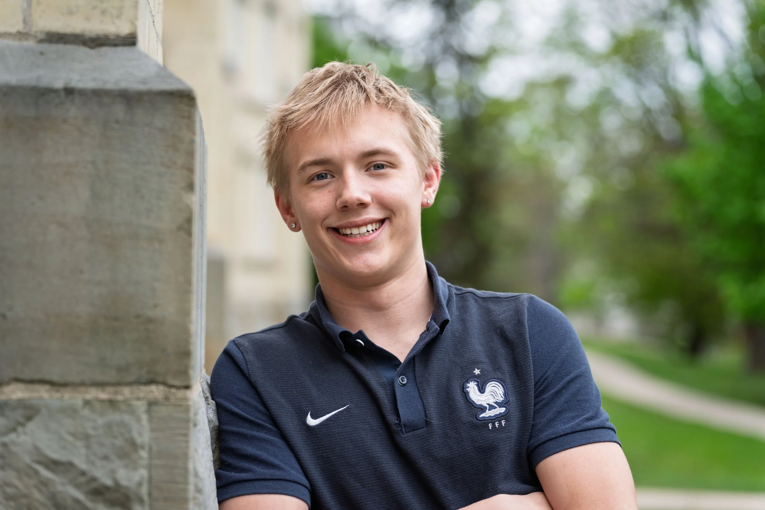 A young man with blond hair smiling outdoors, wearing a navy blue polo shirt with a French national football team logo.