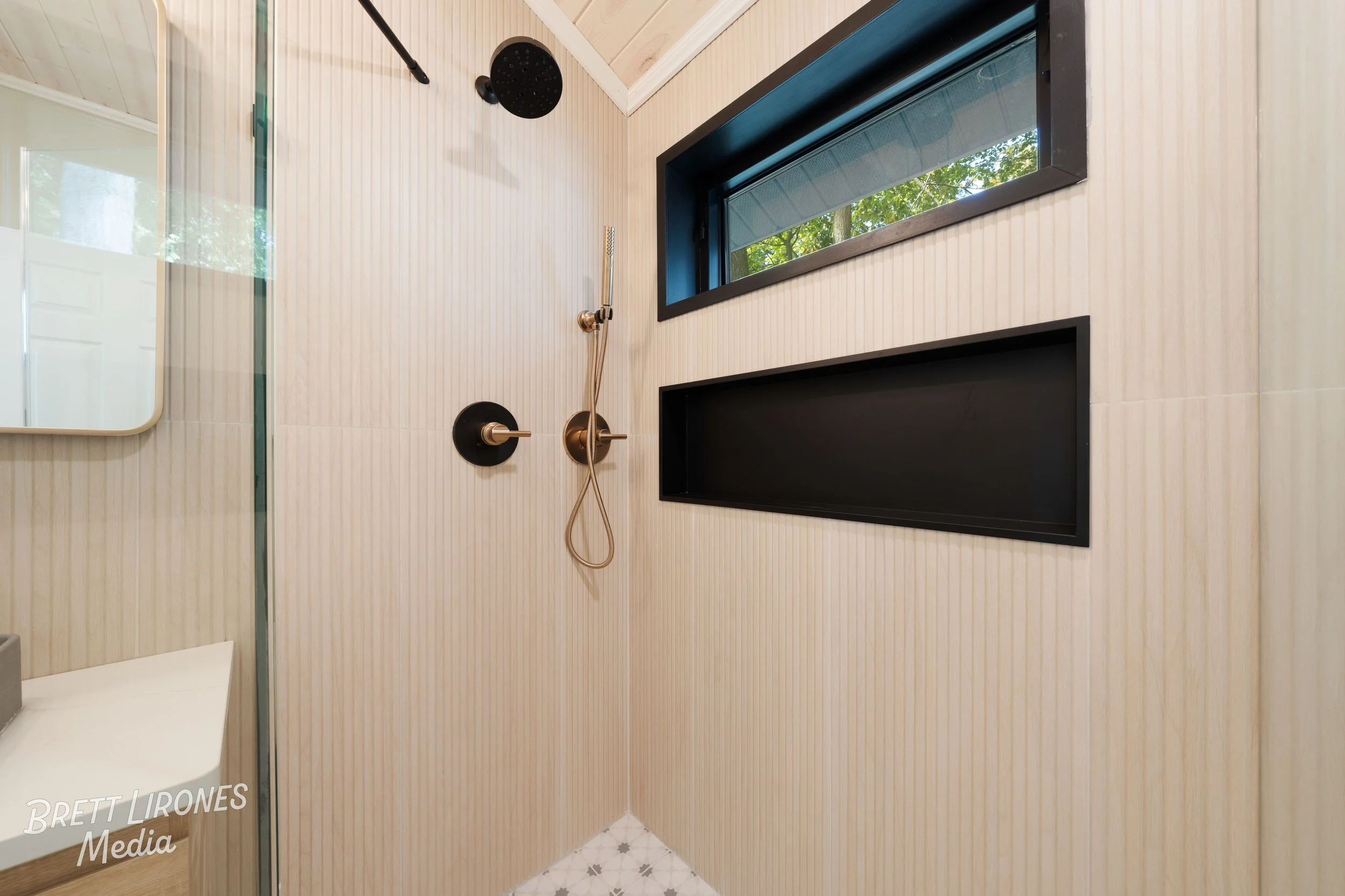 Modern bathroom shower with beige ribbed wall panels, black and brass fixtures, a window showing green foliage outside, and part of a bathroom mirror and sink visible.