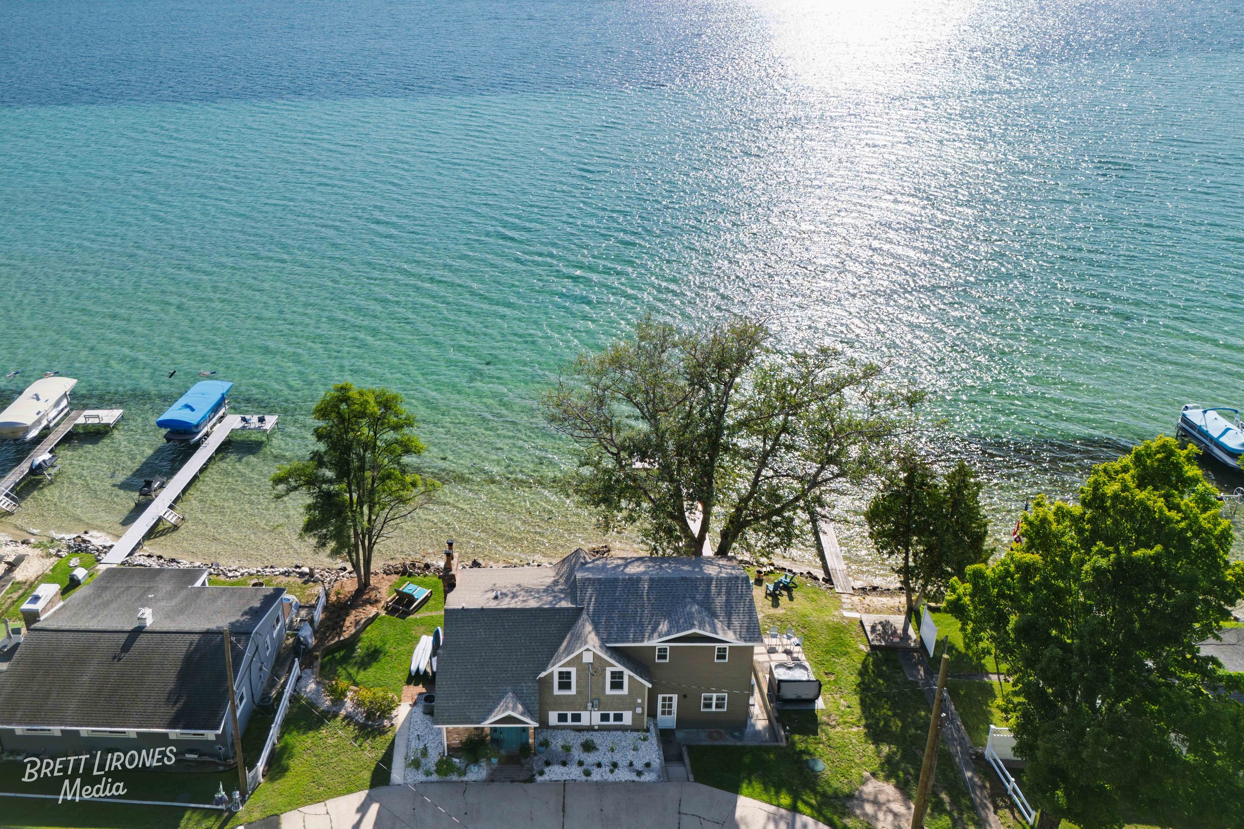 Aerial view of a house by a lake with a small dock, boats, and trees along the shoreline, with sunlight reflecting off the water.