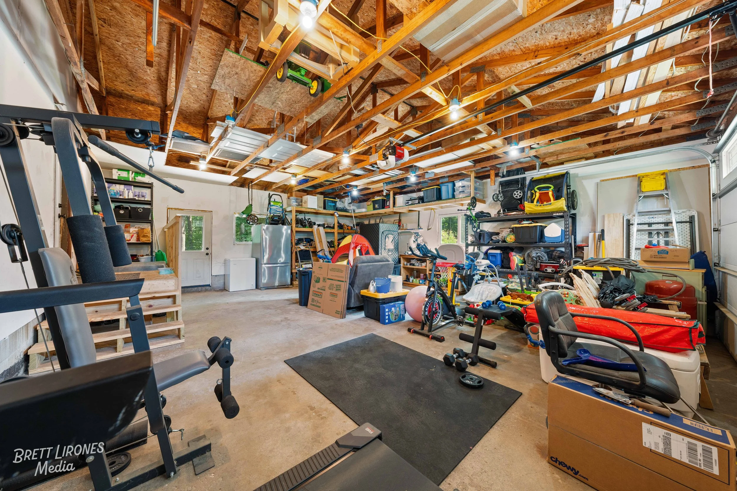 A cluttered garage with an unfinished ceiling and various stored items including workout equipment, toys, boxes, tools, and storage shelves.