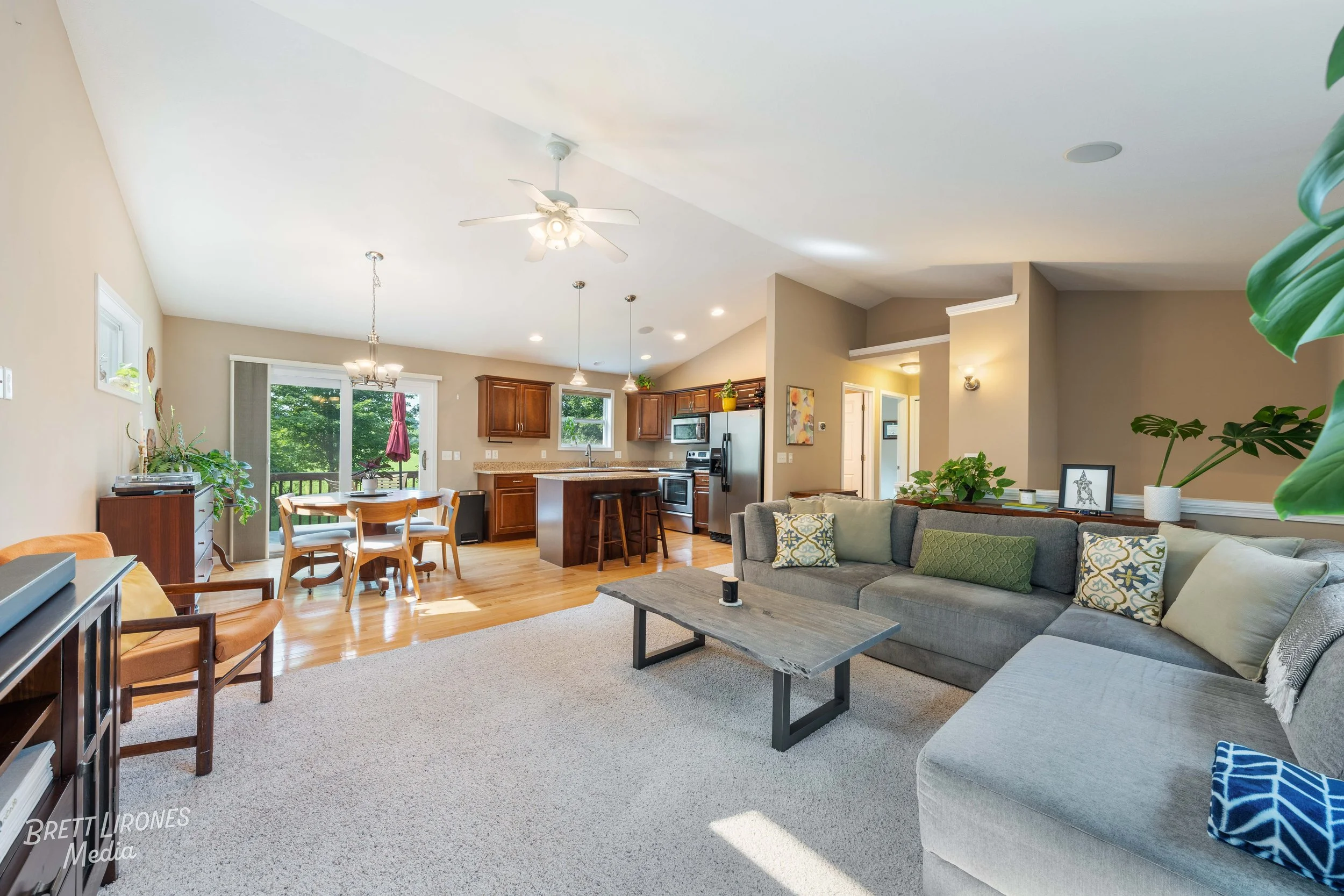 Open-concept living room and kitchen in a modern home with beige walls, a gray sectional sofa with decorative pillows, a wooden coffee table, a dining table with chairs, and stainless steel appliances in the kitchen with wooden cabinets.