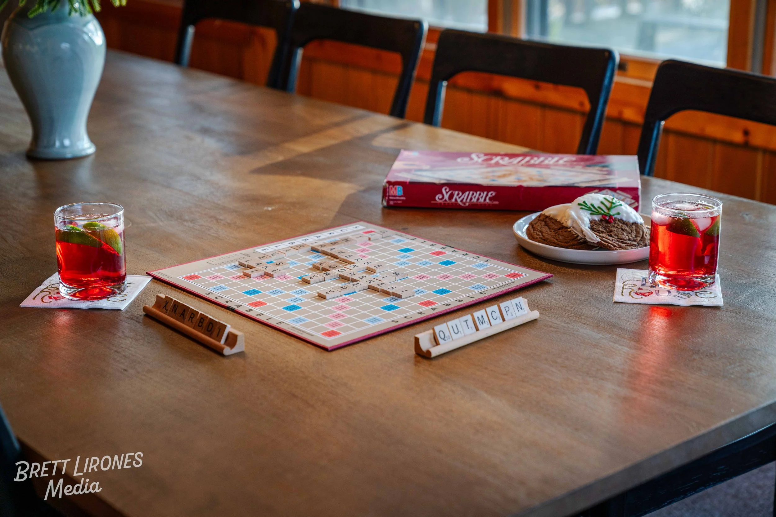 A wooden table set for a game night with Scrabble, two glasses of red drinks with lime slices, a plate with Christmas cookies, and Scrabble game pieces and boards, in a room with wooden walls and windows.