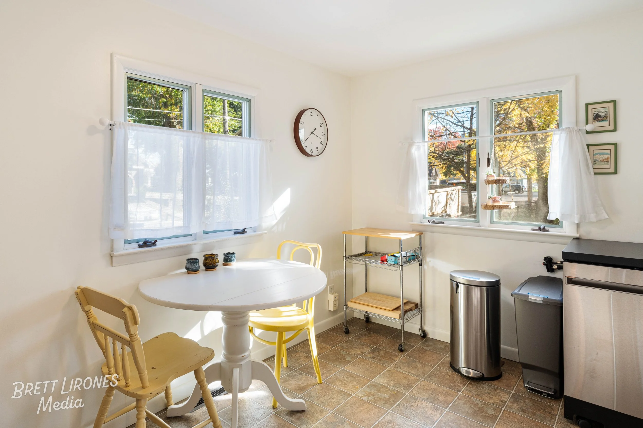 Bright kitchen corner with white walls, three windows with white curtains, a white round table, two yellow chairs, three small candles, a metal shelf with trays and boxes, a wall clock, a trash can, and a stainless steel dishwasher.