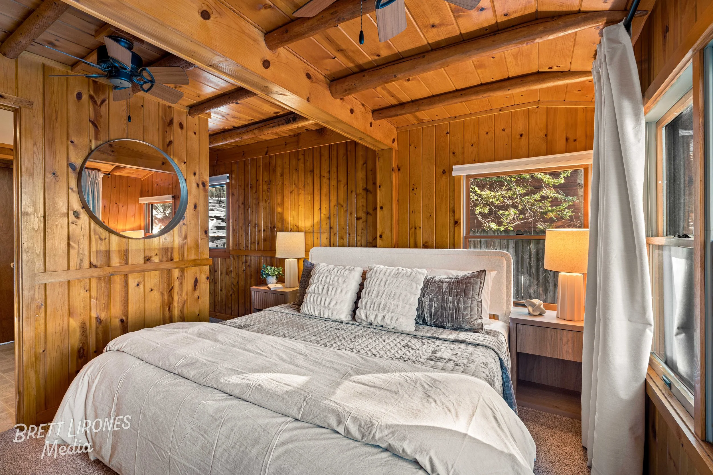 A bedroom with wood-paneled walls and ceiling, a large bed with gray and white bedding, and two bedside tables with lamps, surrounded by windows with curtains, and a mirror on the wall.
