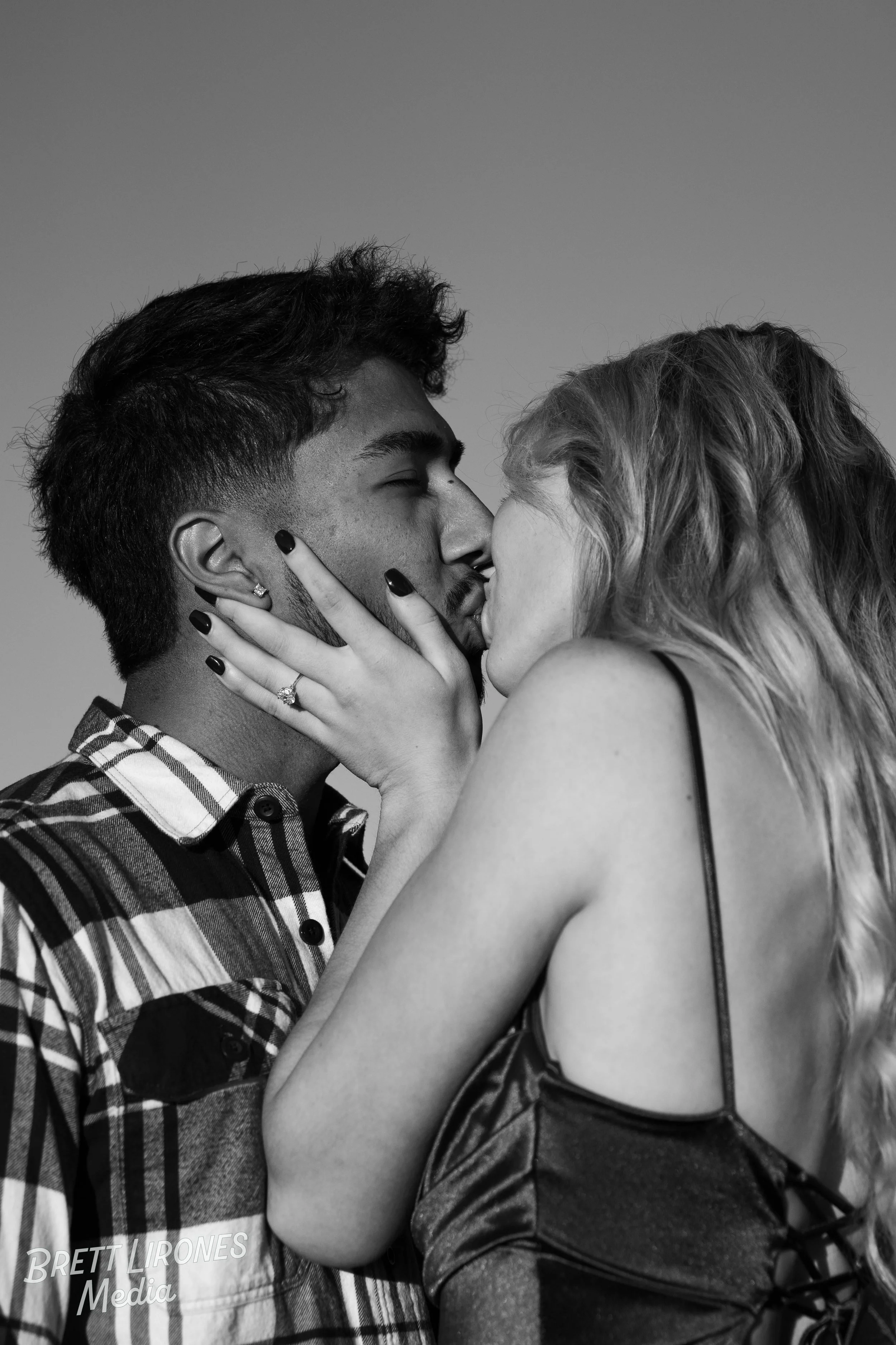 A black and white photo of a young man and woman kissing. The young woman is holding the man's face with one hand, showing an engagement ring. The man has dark hair and a plaid shirt; the woman has long wavy hair and wears a strap camisole.