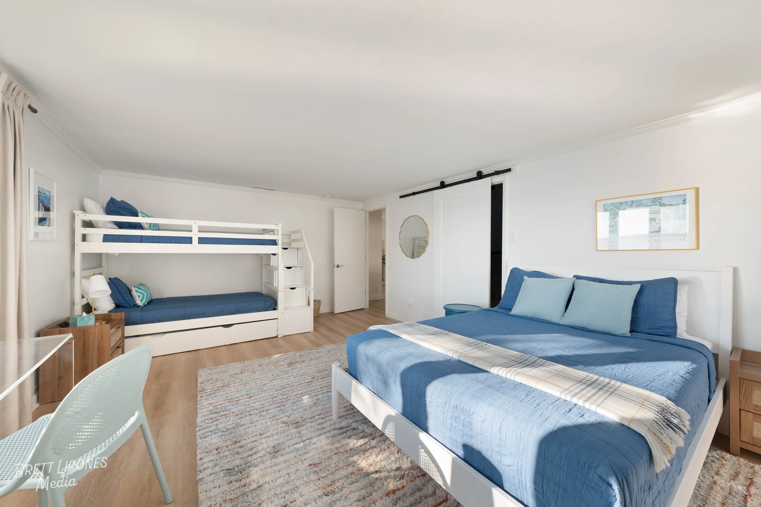 Bright bedroom with white walls and light wood flooring. Features a large bed with blue bedding and pillows, a nightstand on each side, a wooden dresser, and a framed artwork above the bed. A sliding barn door reveals a closet, and a white bunk bed w