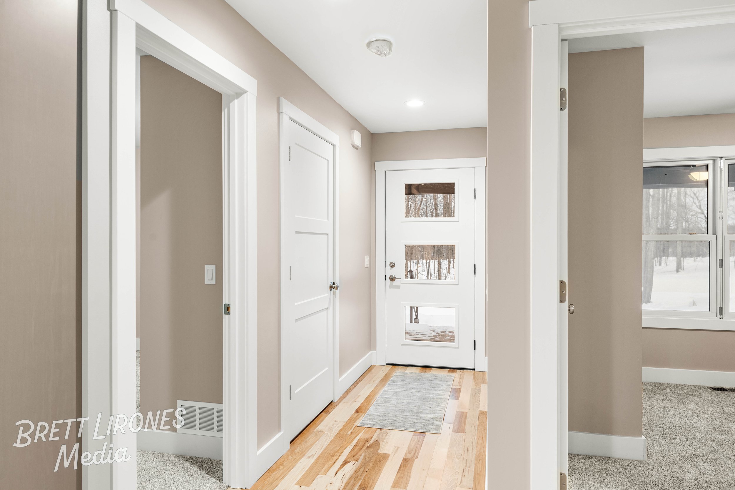 Entryway with hardwood floor, white door with windows, beige walls, carpeted rooms, and a view outside through a window showing snow-covered ground and trees.
