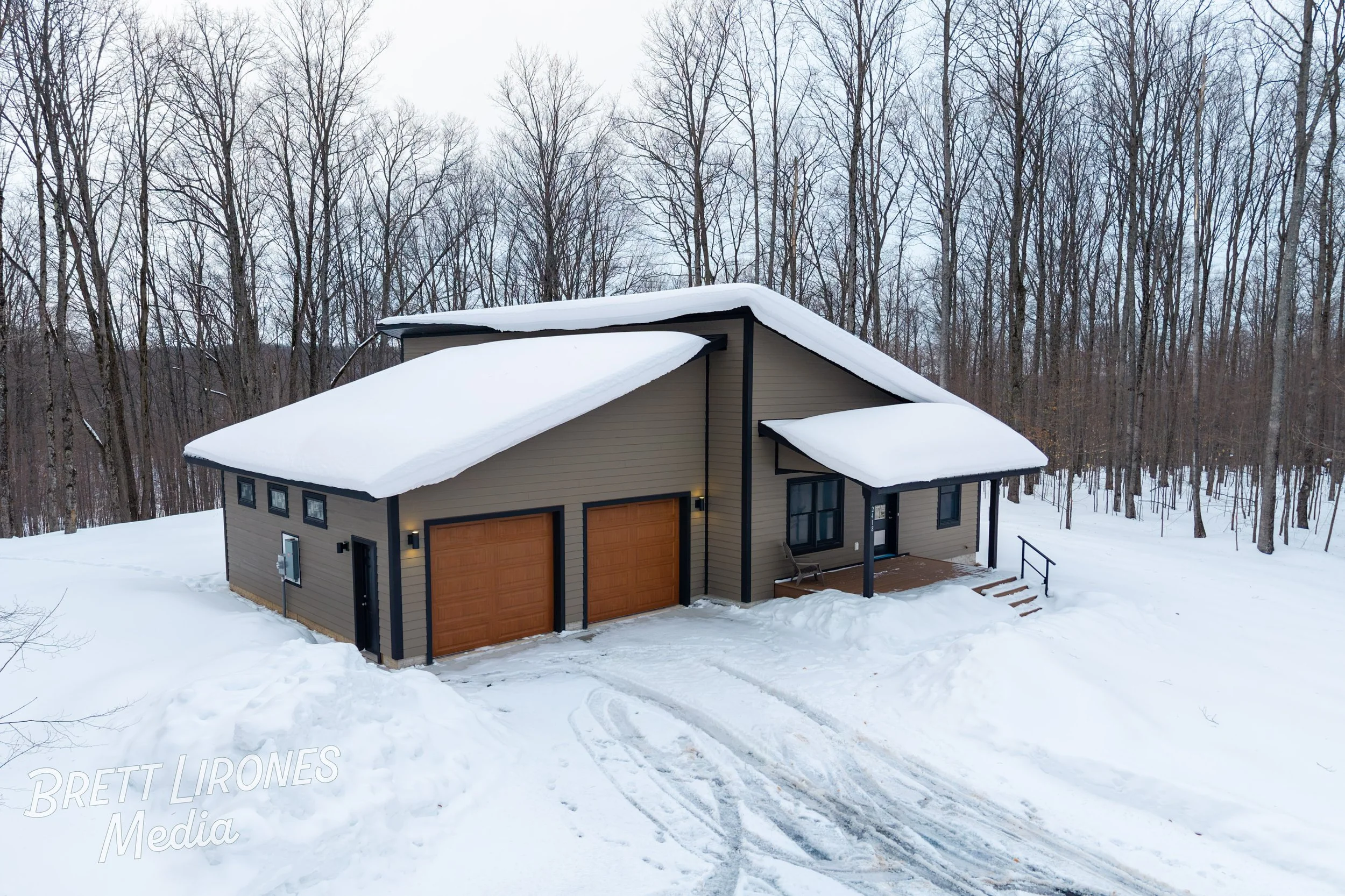 A modern house with a sloped roof covered in snow, situated in a snowy landscape with leafless trees in the background. The house has double garage doors, a small front porch with stairs, and a driveway with visible tire tracks.