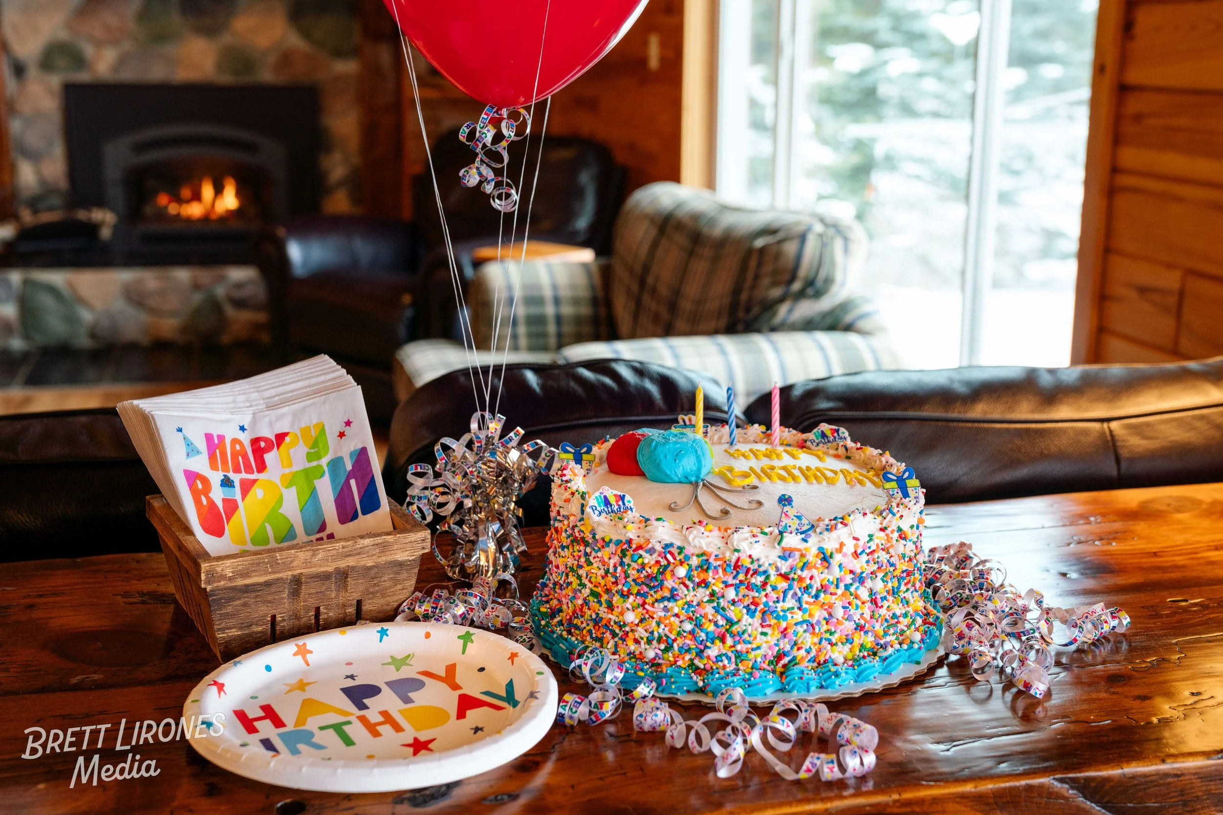 Colorful birthday cake decorated with rainbow sprinkles, topped with two balloons, candles, and birthday messages, surrounded by celebratory plates and party accessories on a wooden table inside a cozy cabin with a fireplace and large windows.