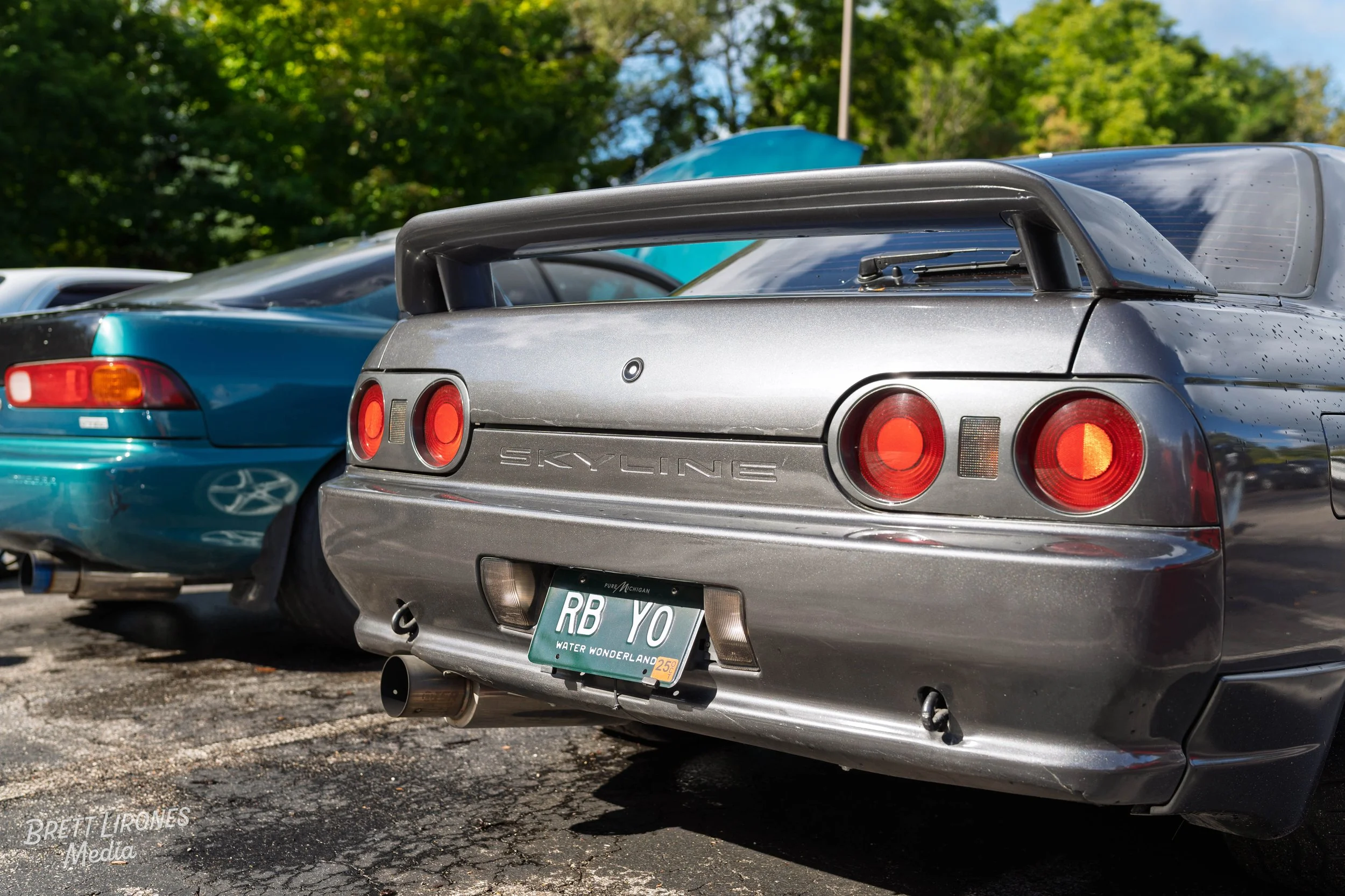Rear view of a gray Nissan Skyline sports car parked in a lot, with a blue car next to it, in a natural outdoor setting with green trees.
